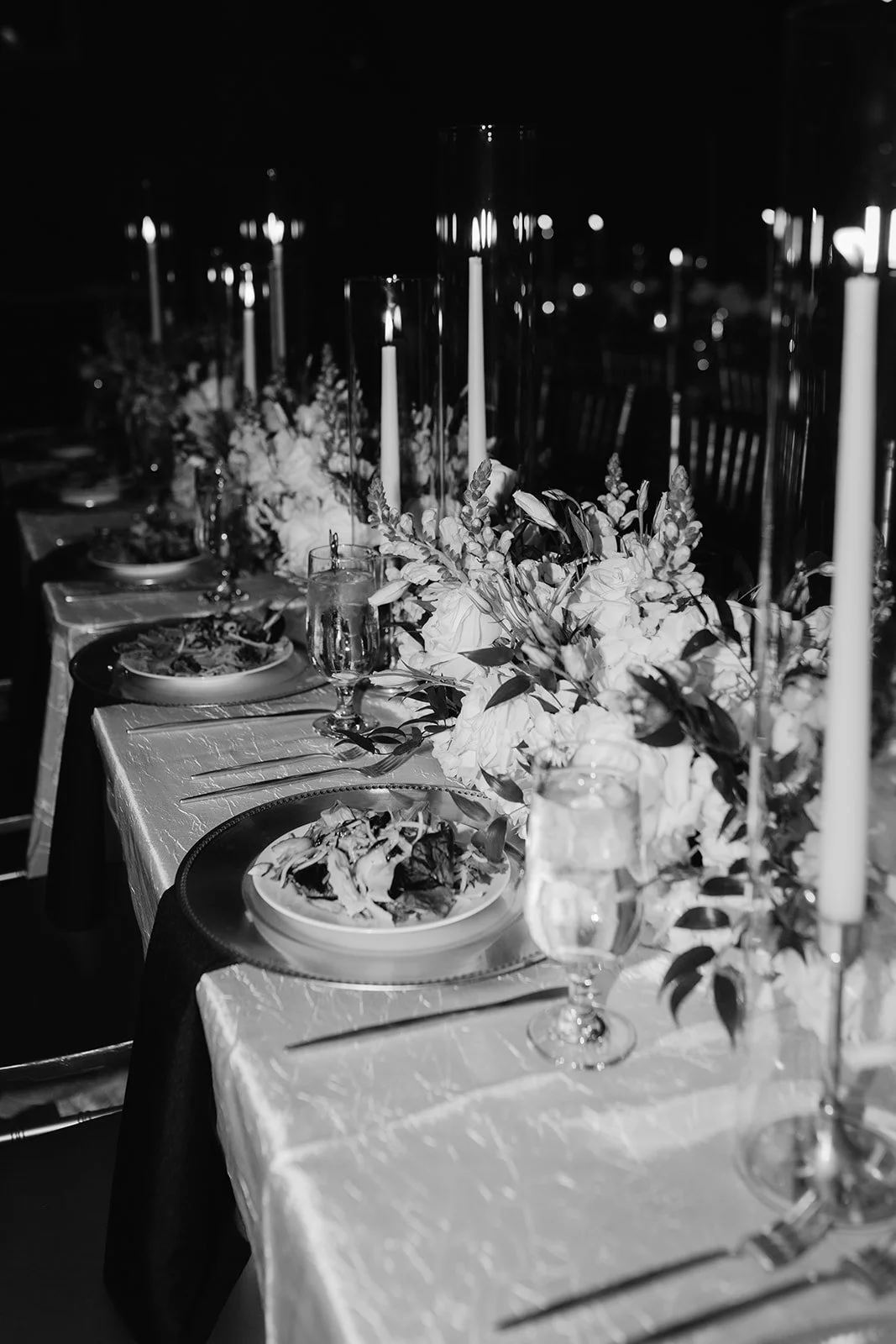 A elegantly set dinner table with candles, floral centerpieces, plates of salad, wine glasses, and a tablecloth, likely for a special occasion or formal event.