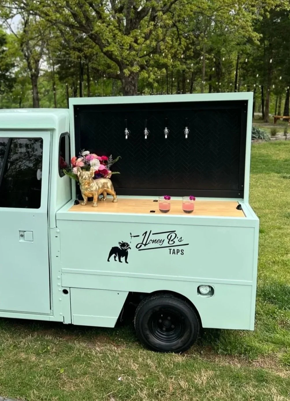 An ice cream or beverage stand on a light green truck with the name 'Honey B's Taps' and a black bear logo. The stand features four taps, a small floral arrangement, and two pink drinks with flowers on top. There are greenery and trees in the background.