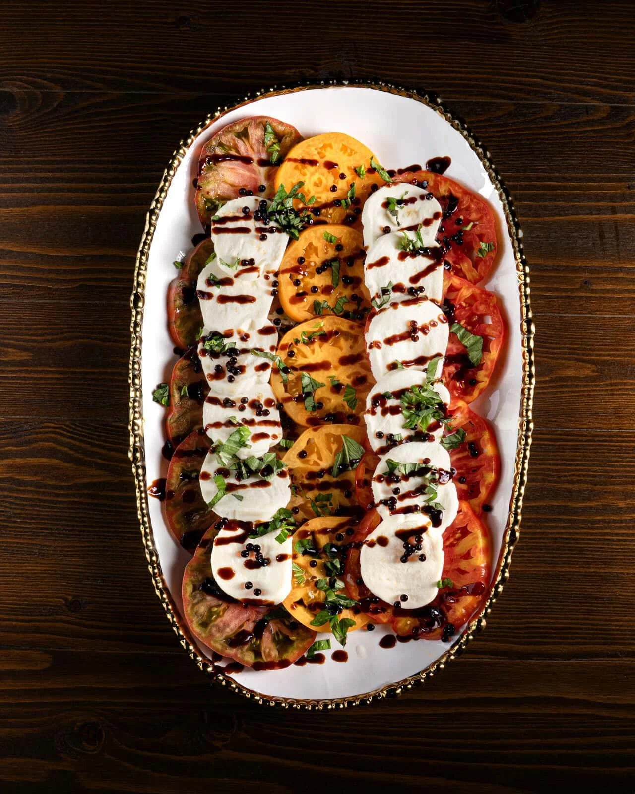 A platter of heirloom tomato salad with slices of mozzarella cheese, chopped herbs, balsamic glaze, and black pepper on a white oval dish with a decorative gold rim.