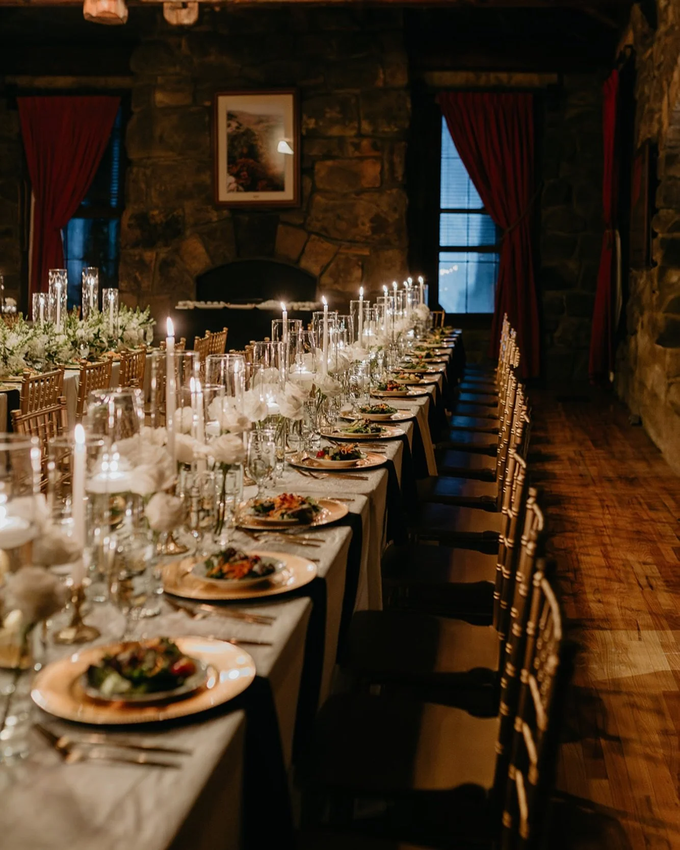 A long dining table set for a formal dinner with plates, silverware, wine glasses, and floral centerpieces, inside a rustic stone-walled room with windows and red curtains.