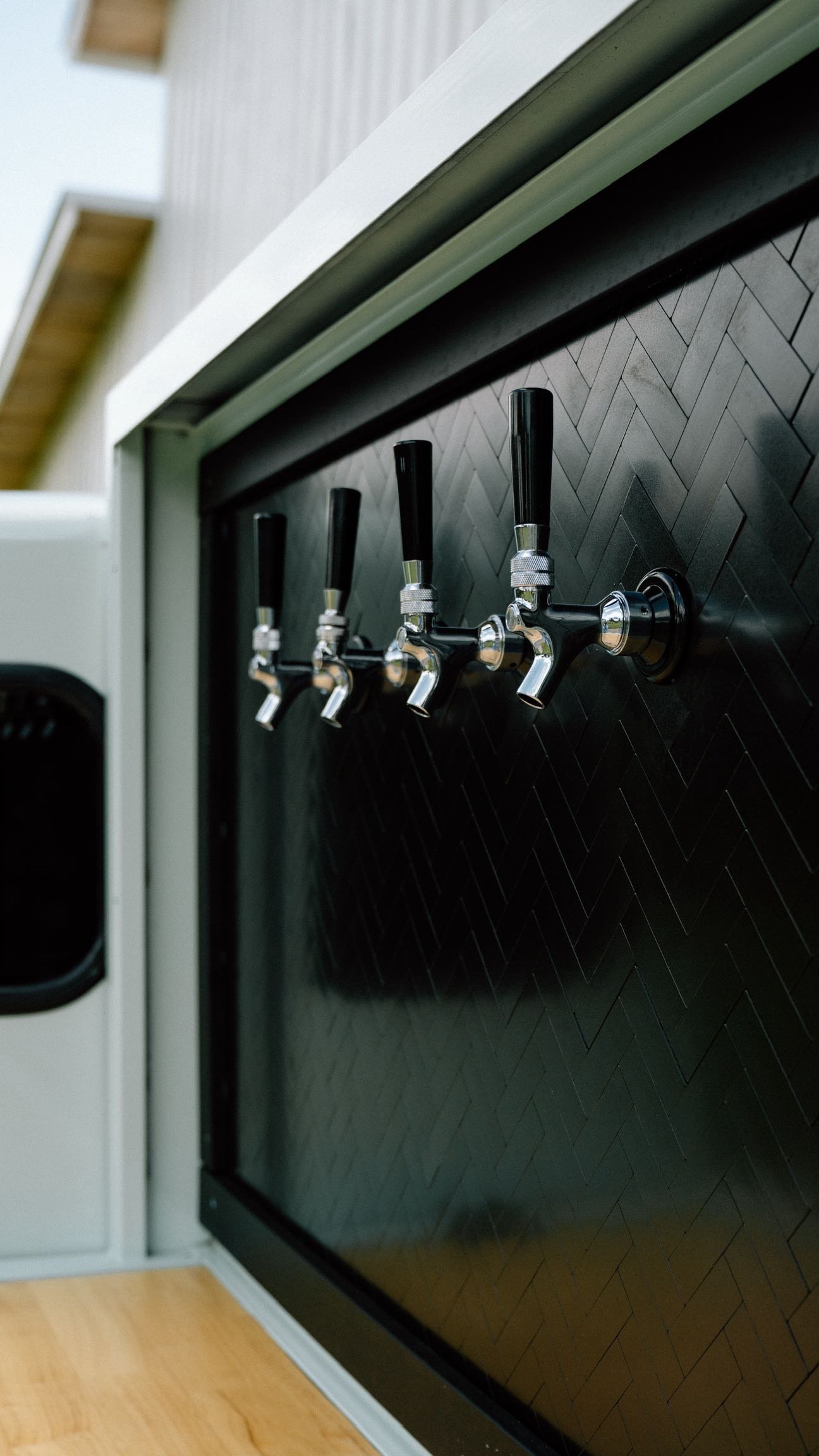 A set of five black and silver beer taps mounted on a black herringbone-patterned wall.