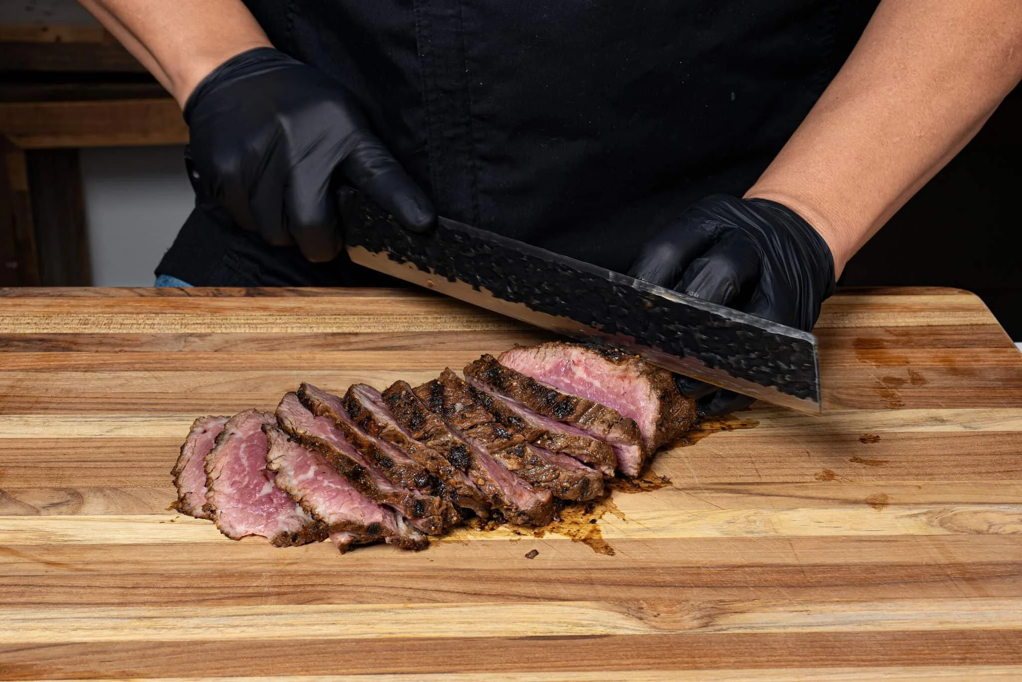 Person slicing a cooked steak on a wooden cutting board, wearing black gloves.