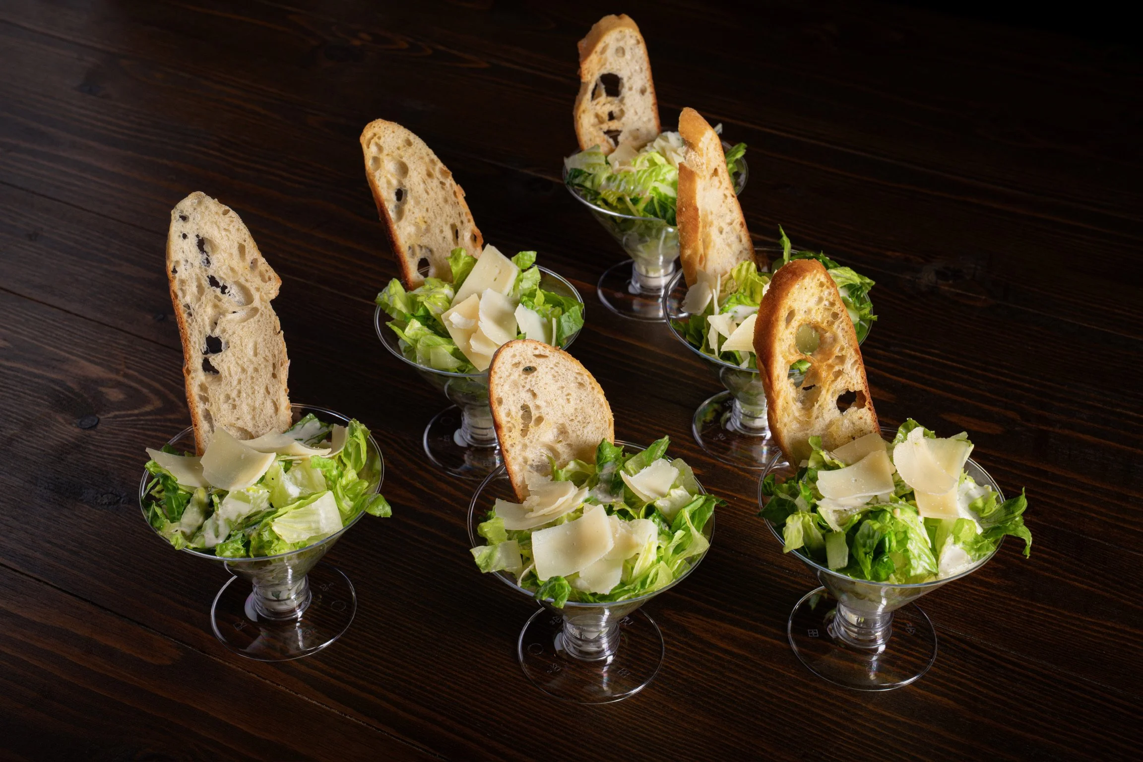 Six small glass bowls filled with chopped lettuce, shaved cheese, and a crispy breadstick standing upright in each bowl on a dark wooden table.