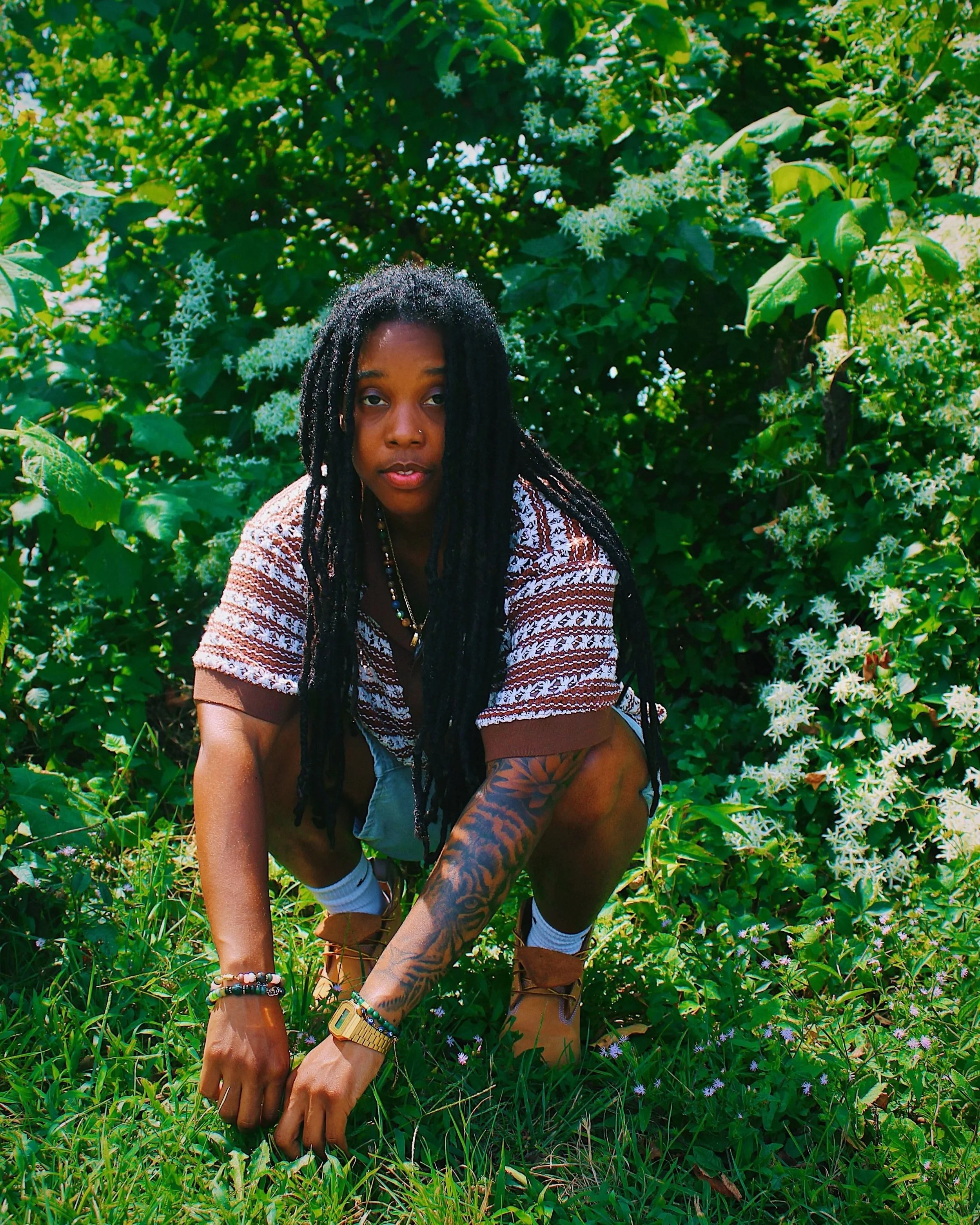 A person crouching in a lush green garden, surrounded by various plants and white flowers, wearing a patterned shirt, shorts, and boots.