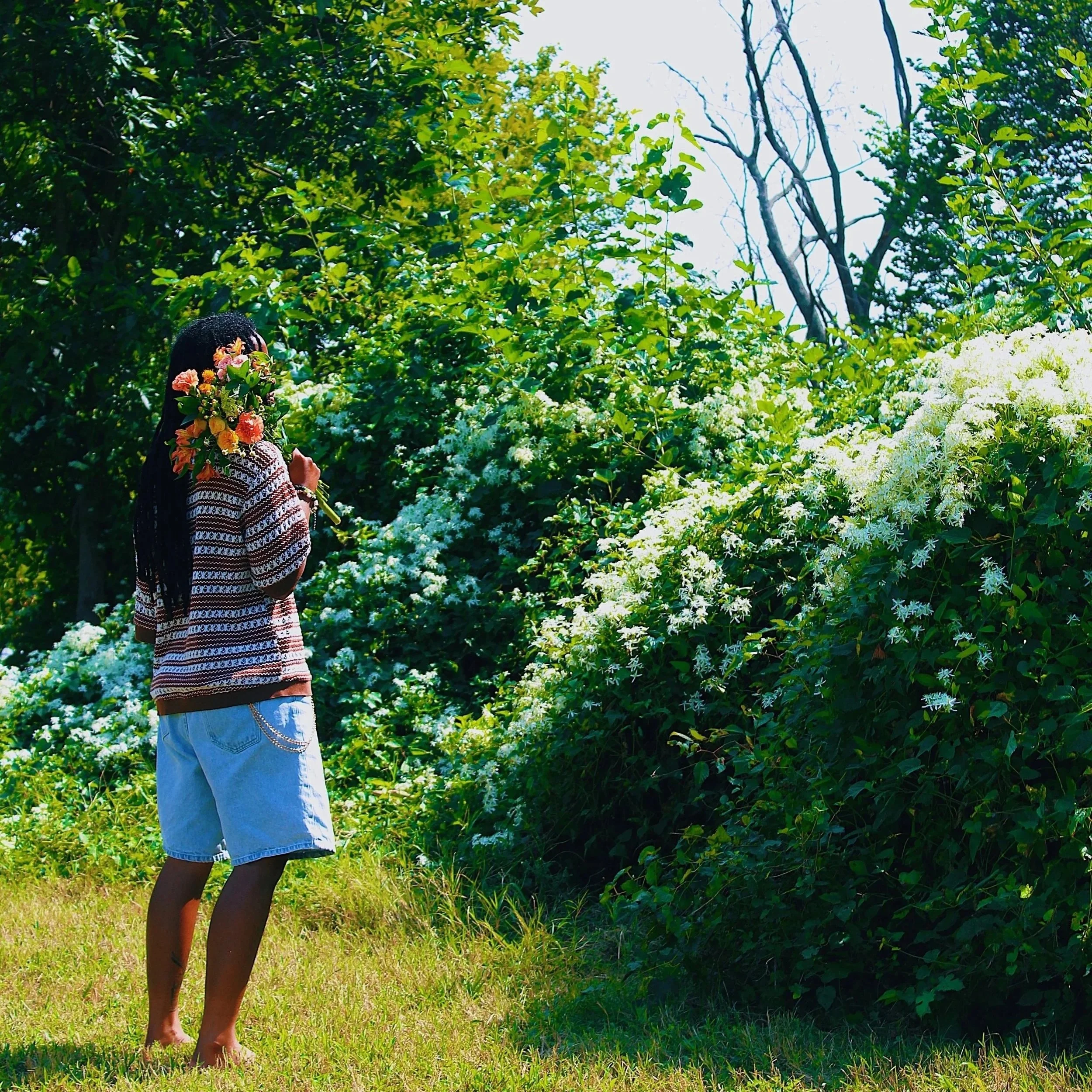 Person with long hair holding a bouquet of flowers, standing on grass near bushes with white flowers, surrounded by trees, facing away from the camera.