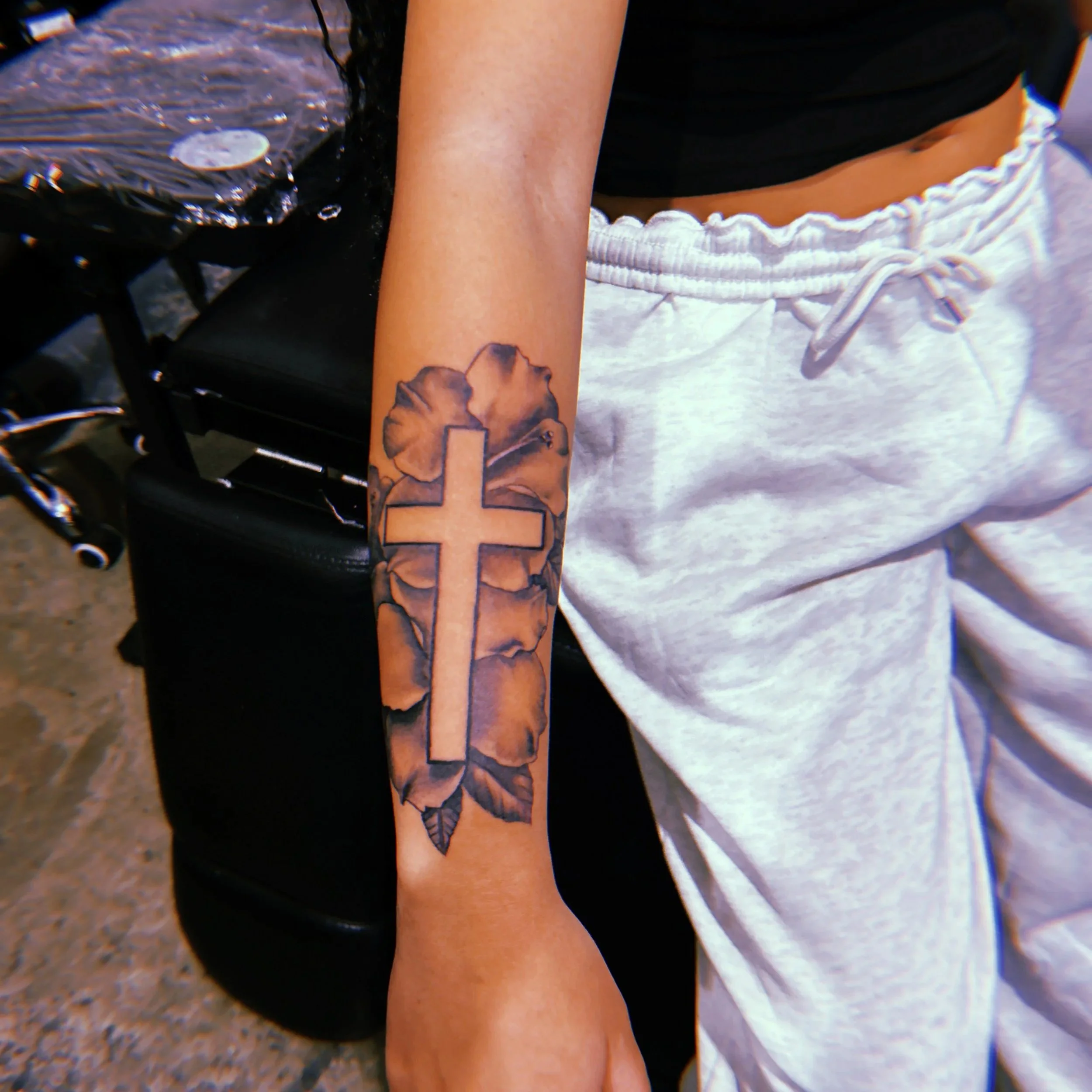 A person's arm with a tattoo of a cross on a flower with leaves on their forearm.