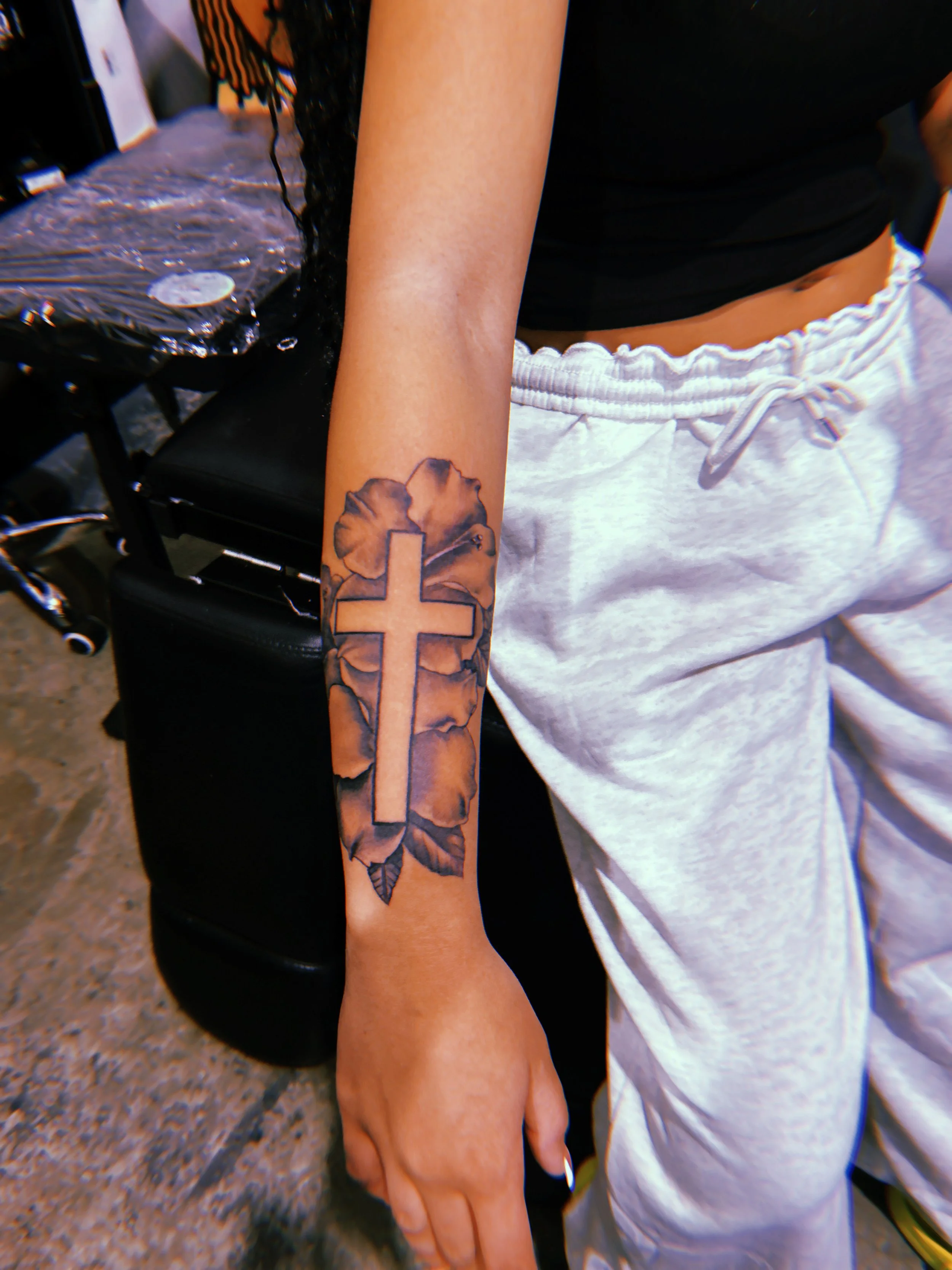 A person with a tattoo of a cross on their forearm, featuring black and gray shading and leaf details, wearing a black top and grey sweatpants.