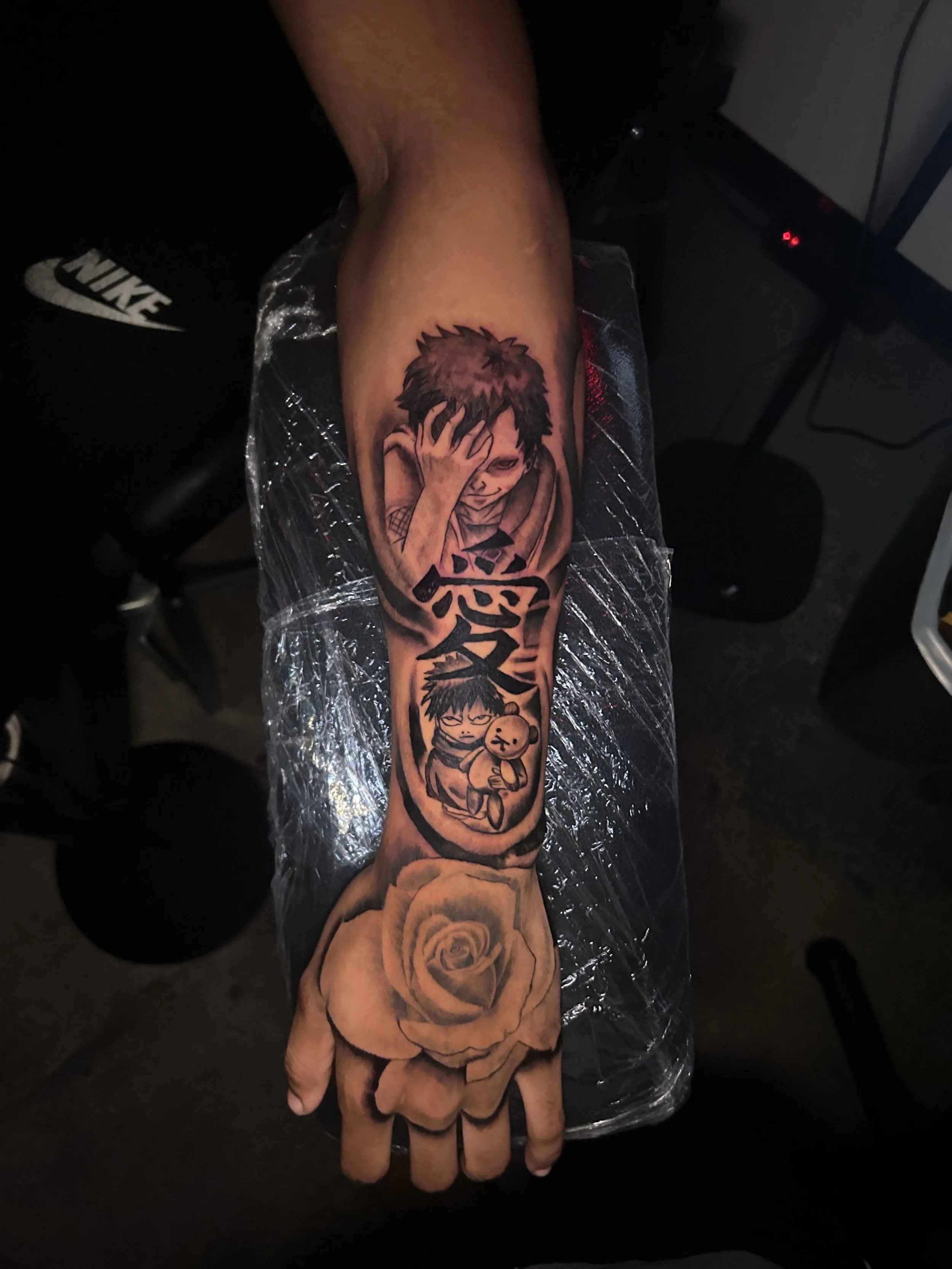 Tattoo of two anime-style characters with one covering his eye and a teddy bear, along with a large rose at the wrist.