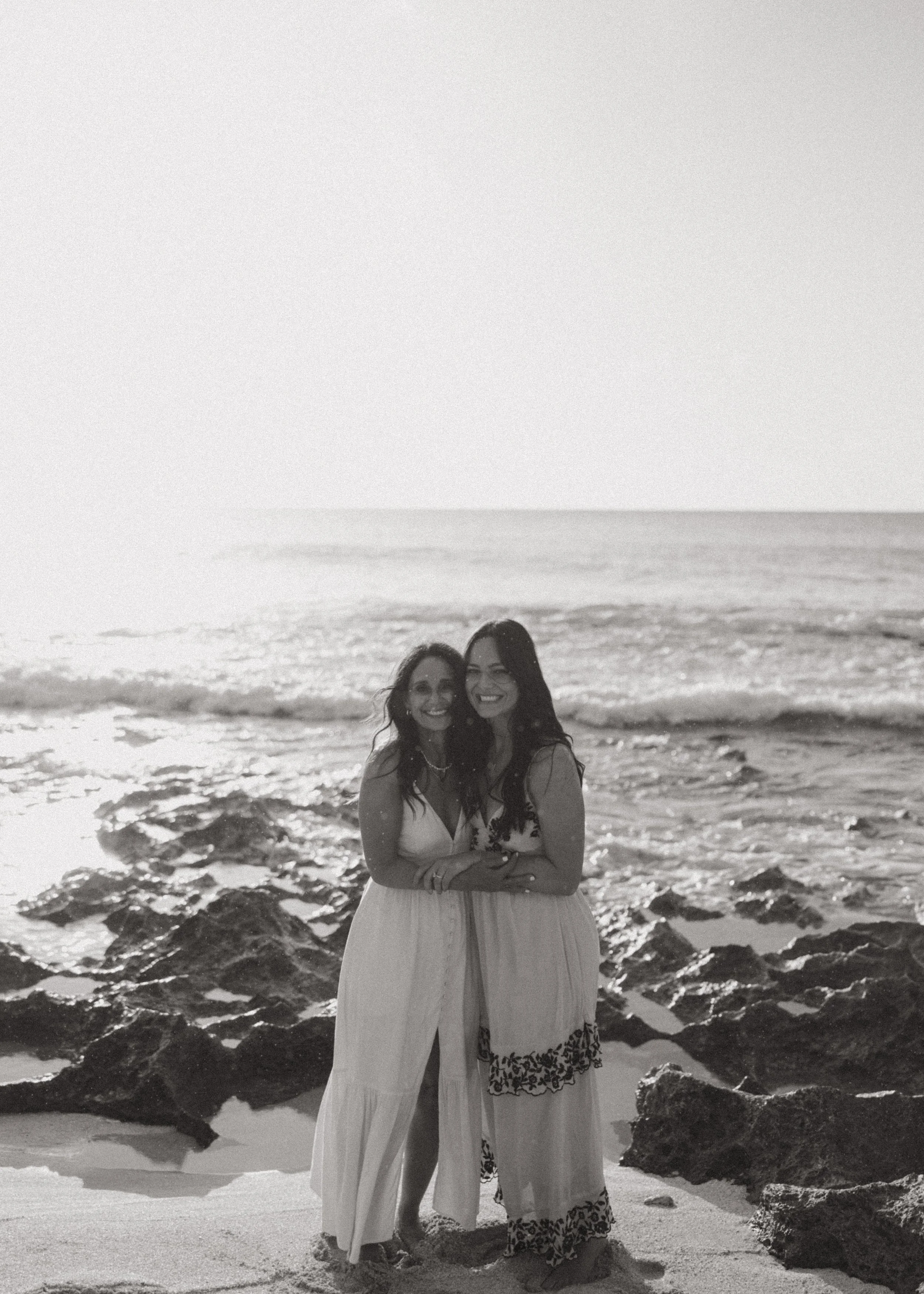 Two women standing close together on a beach with rocks and the ocean in the background, smiling at the camera.