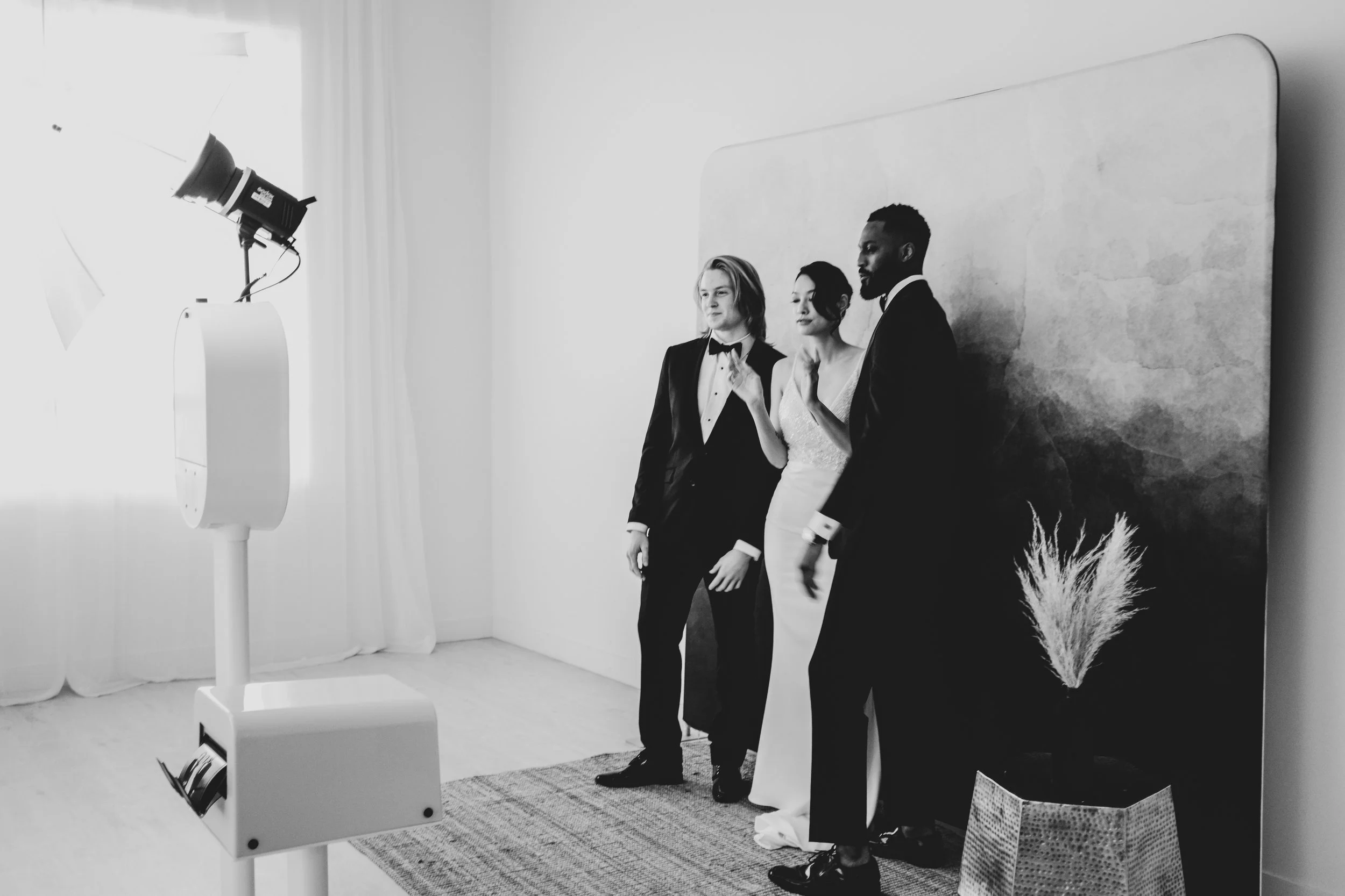 Black and white photo of three people dressed in formal attire, standing in front of a painted backdrop with a potted plant nearby. They appear to be posing for a photo or participating in a photoshoot.