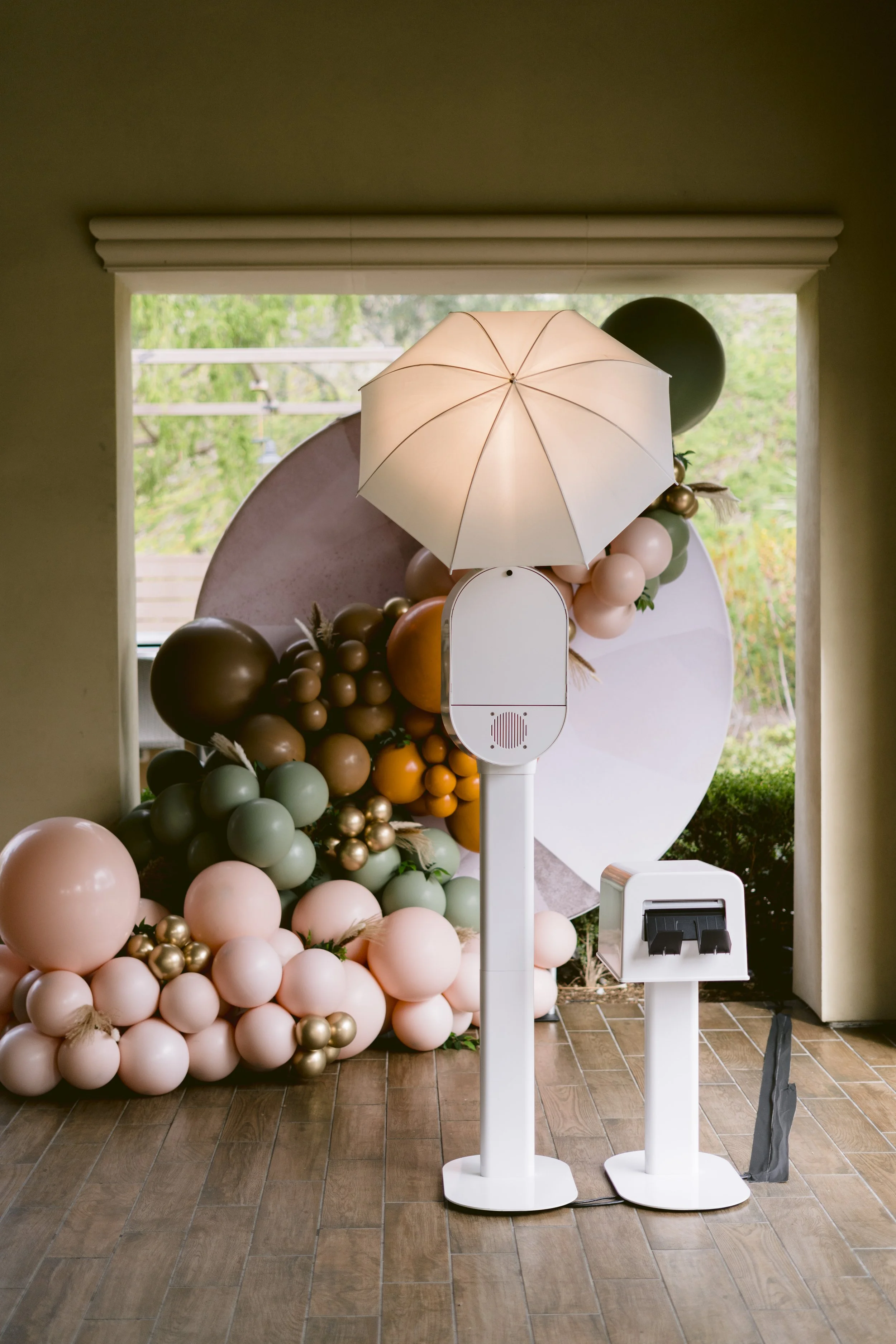 Indoor balloon decoration display with pastel and metallic balloons, a standing umbrella, and photo booth props on a wooden floor.
