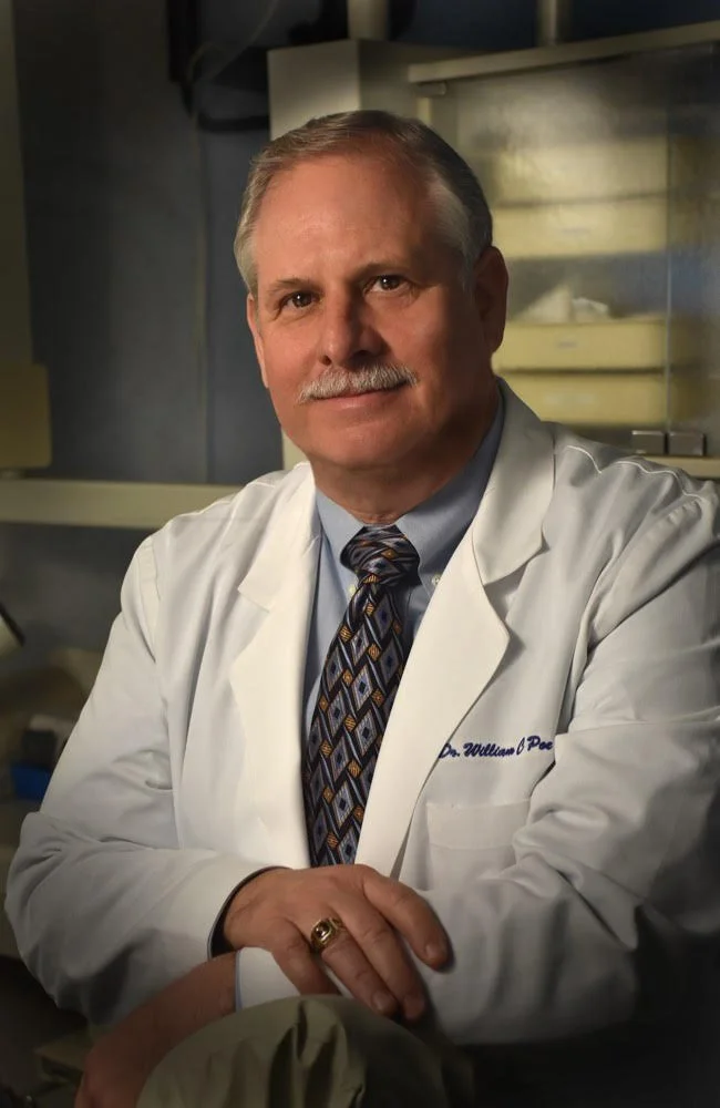 A middle-aged man with a mustache wearing a white lab coat, light blue dress shirt, and patterned tie, sitting in a medical or laboratory setting.