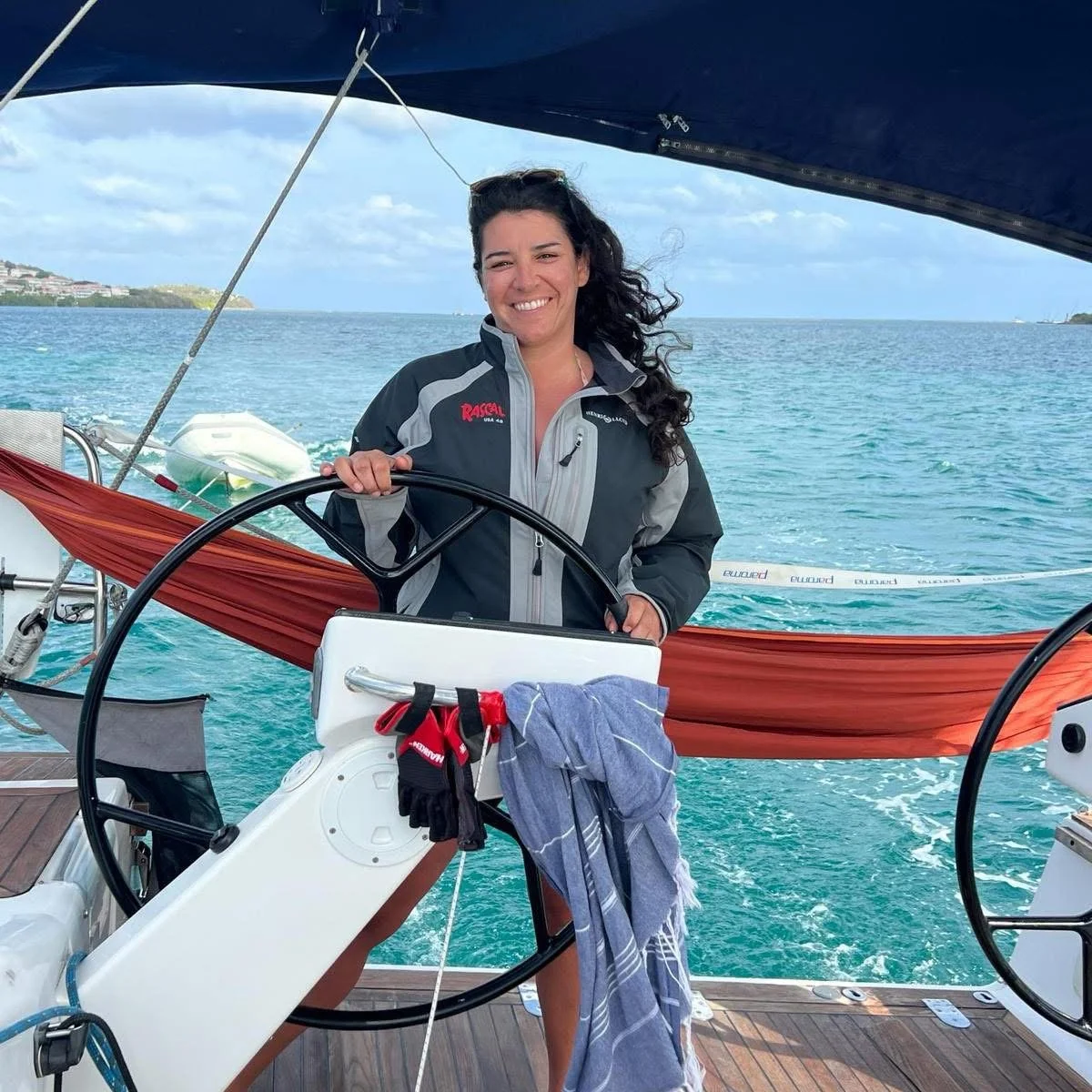 A smiling woman wearing a jacket standing at the helm of a sailboat with ocean and cloudy sky in the background.
