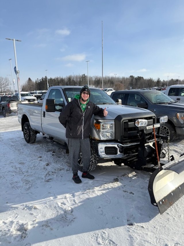 A person standing next to a pickup truck with a snowplow attachment in a snowy parking lot, giving a thumbs-up.