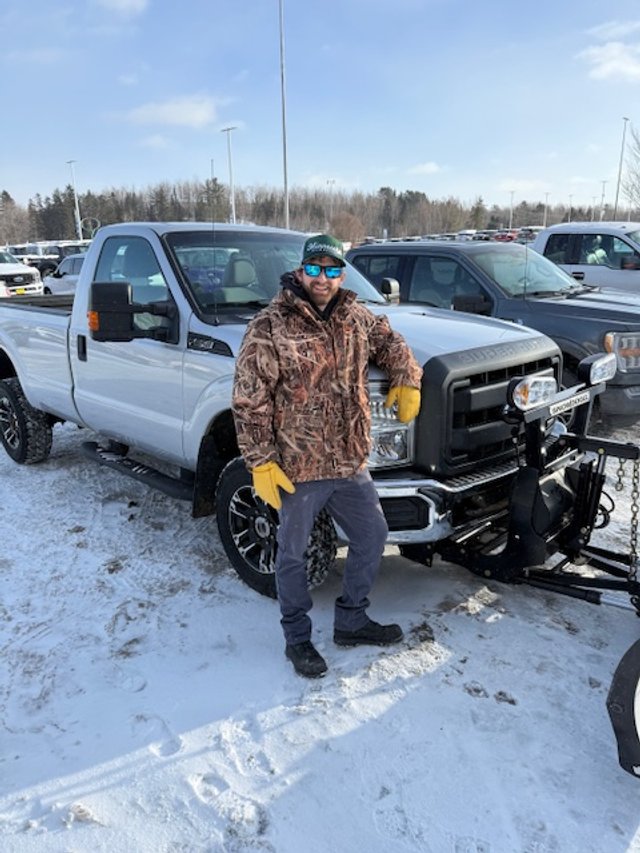 A man in a brown camo jacket, blue jeans, yellow gloves, sunglasses, and a baseball cap standing next to a white pickup truck with a plow in a snow-covered parking lot.