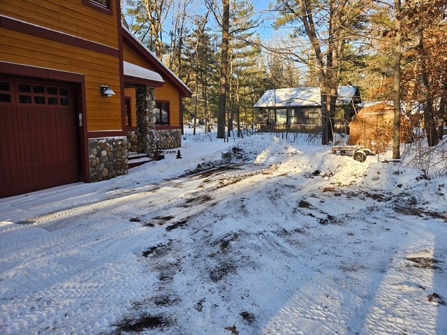 A snow-covered driveway in front of a wooden house with a garage on the left and another house visible in the background amidst trees.