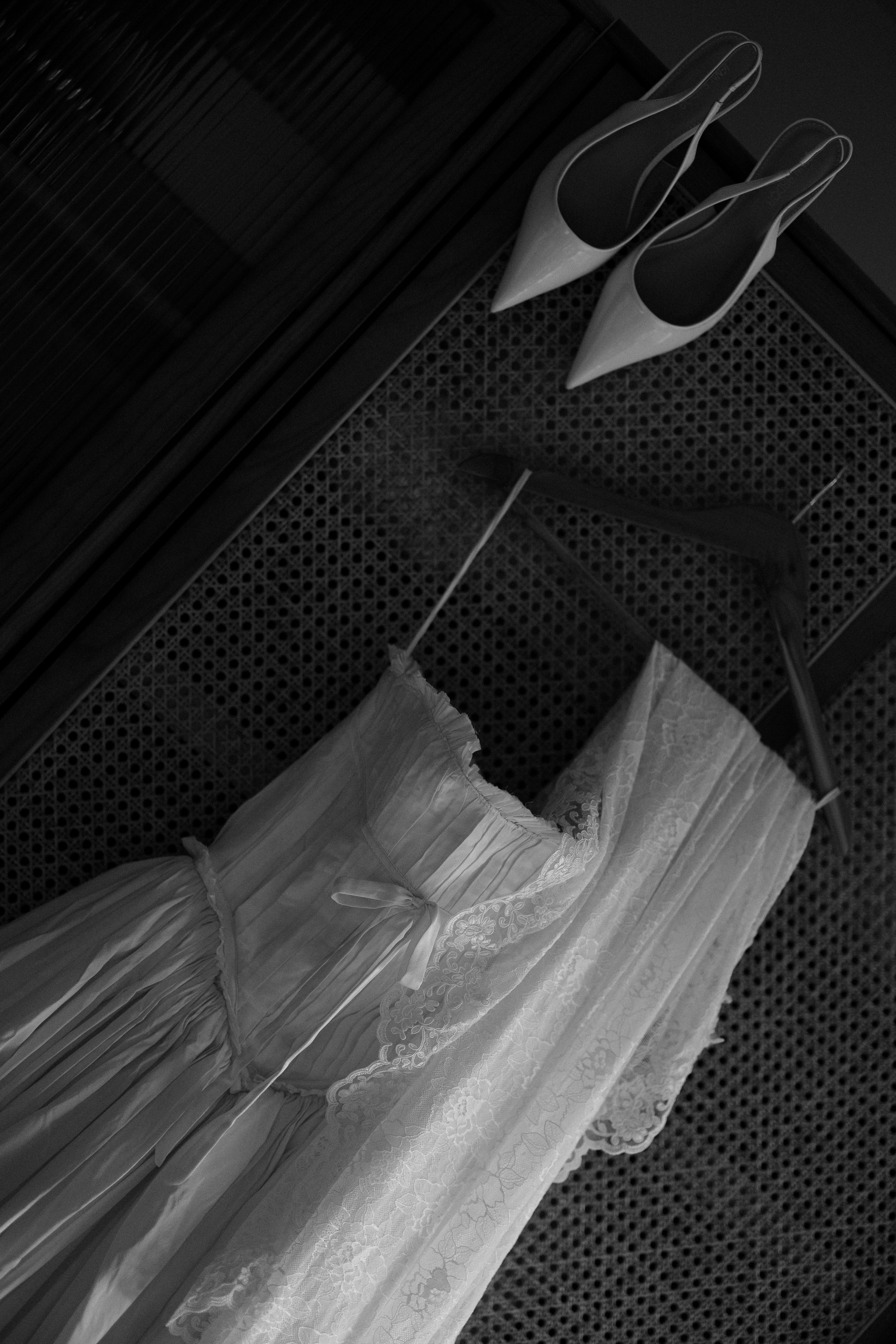 Black and white photo of a wedding dress hanging, accompanied by white high heels placed on a lace-covered surface. Taken by wedding videographer NYC.