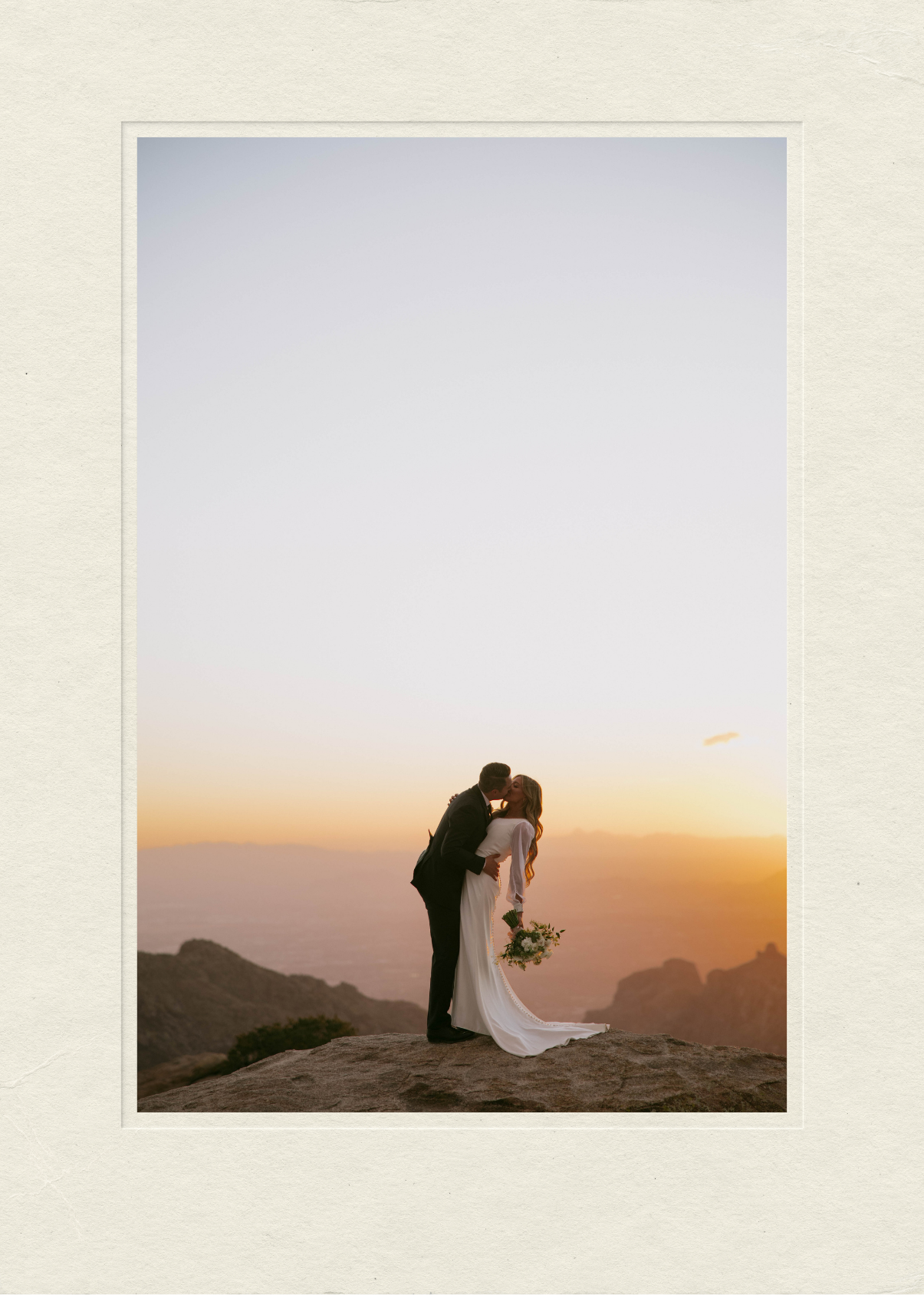 A couple in wedding attire kisses on a mountaintop during sunset, with mountains and sky in the background. Documented by Arizona wedding videographer.