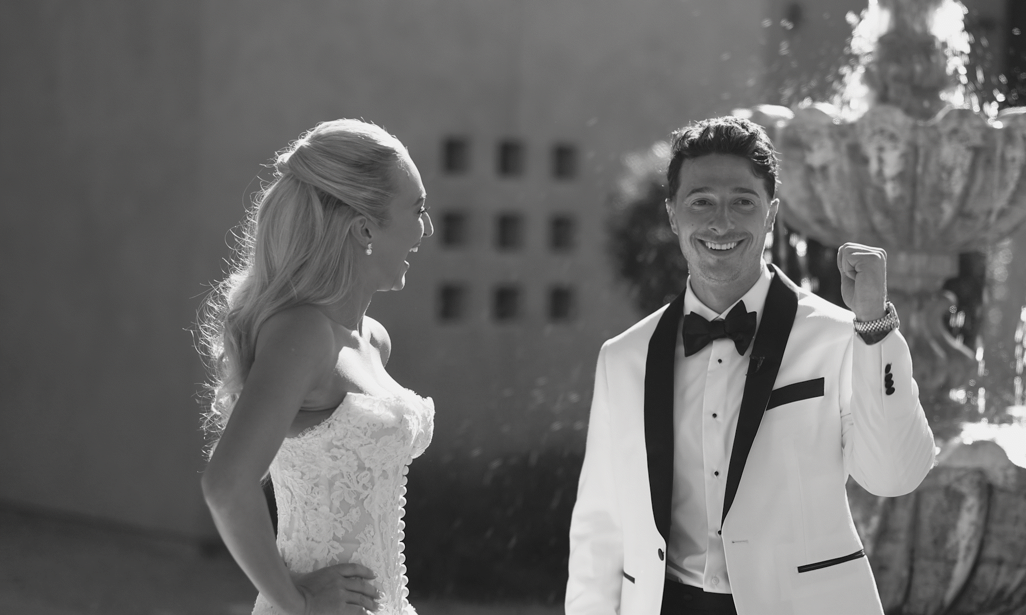 A newlywed couple in wedding attire celebrating outdoors. The woman is laughing while looking at the man, who is smiling and clapping, recorded by wedding videographer Arizona.