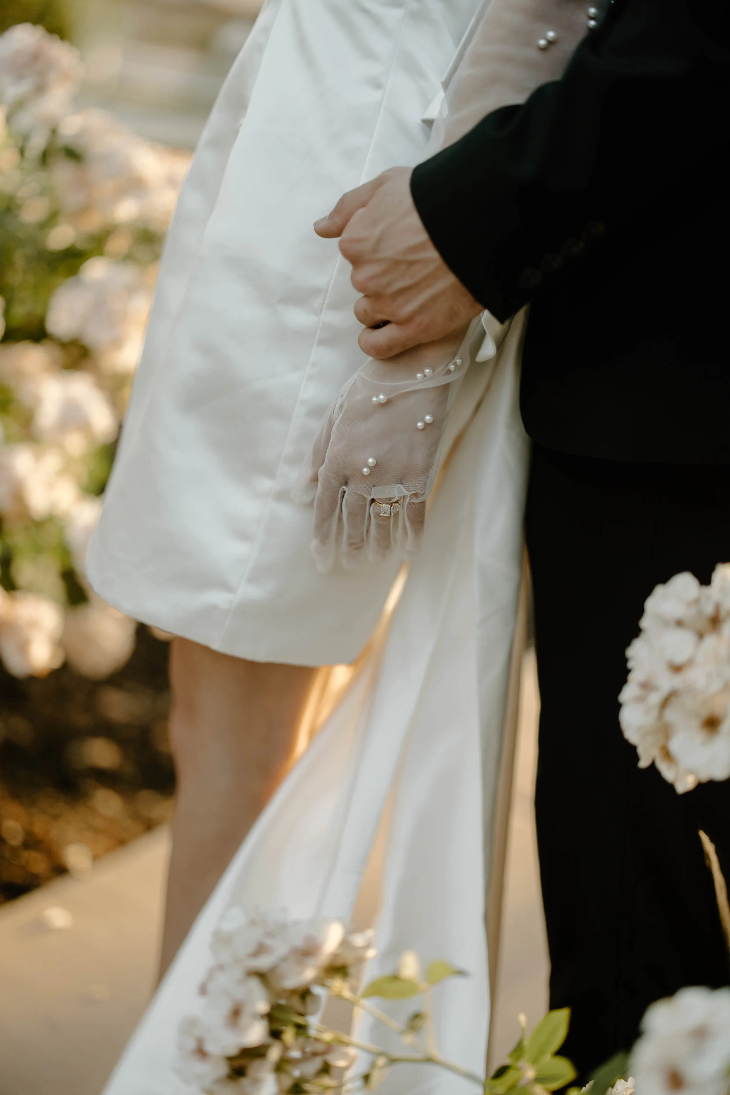 Close-up of a bride and groom holding hands during a wedding ceremony, focusing on the bride's gloved hand with pearl details and the groom's hand, with floral decorations in the background. Captured by destination documentary videographer.