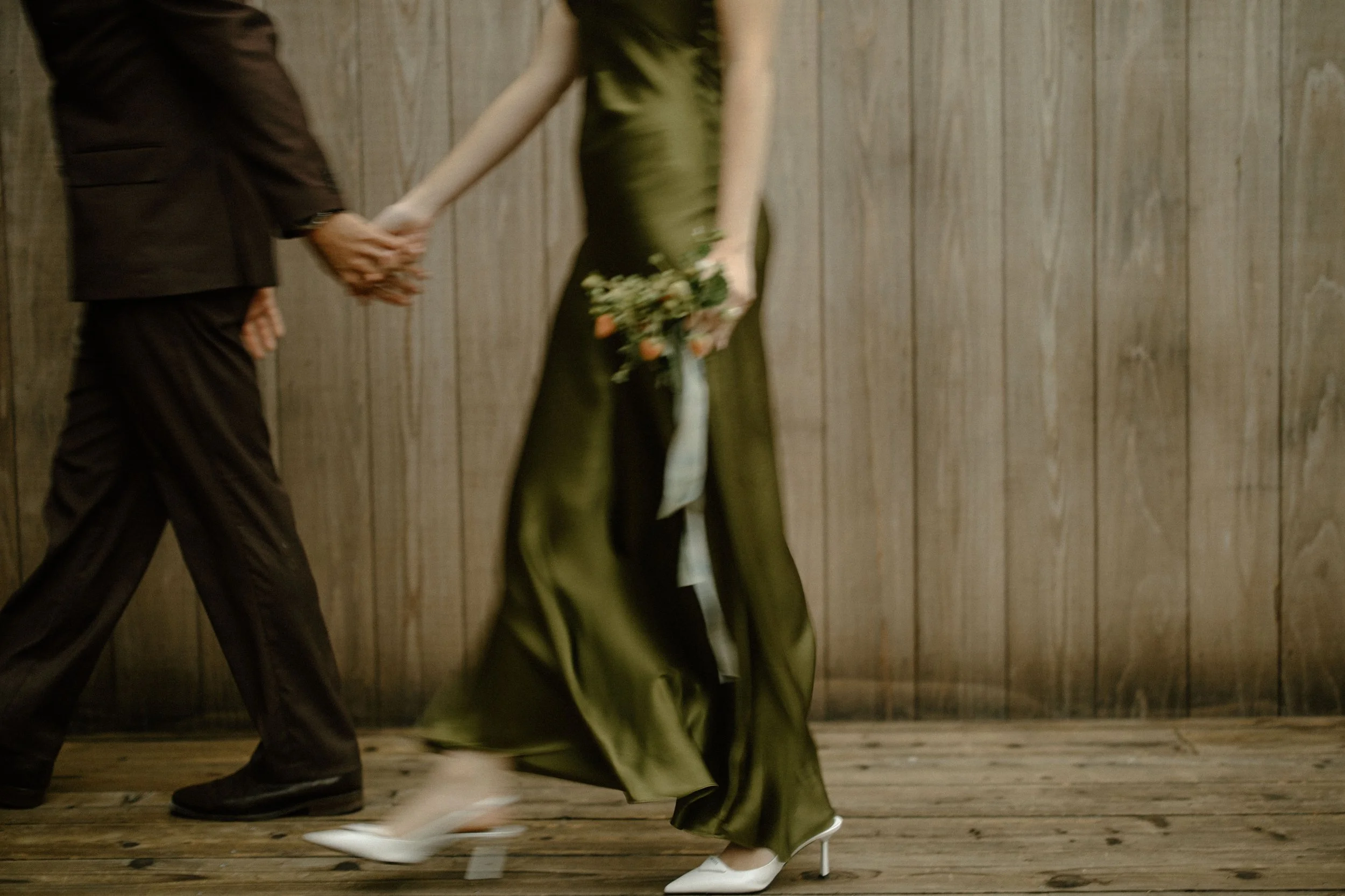 A woman in a long green dress holding a bouquet of flowers, walking hand-in-hand with a man in dark clothing, against a wooden wall background. Image taken by destination film videographer.