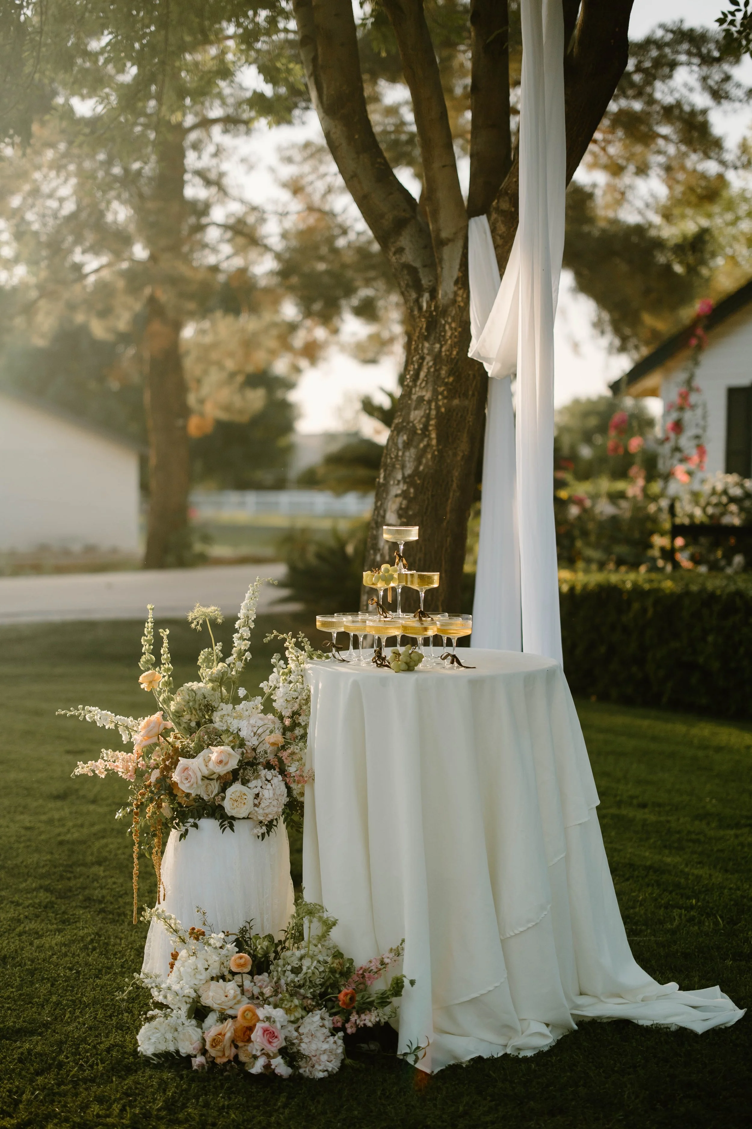 Outdoor wedding setup with a champagne tower on a round white table, decorated with a large floral arrangement of white and pink flowers, with white fabric draped around a tree in the background during sunset. By destination film videographer.