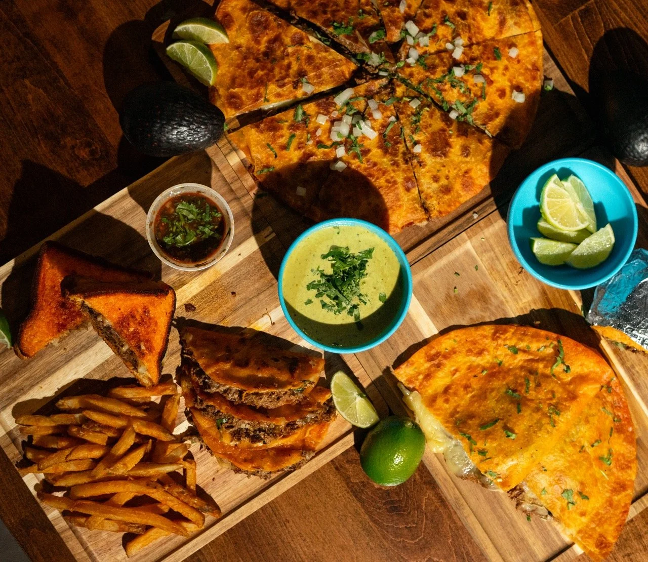 A wooden table with two wooden serving boards holding various Mexican dishes. The top board has two slices of cheesy pizza with chopped onions and cilantro, lime wedges, and avocados. The bottom board has French fries, a slice of garlic bread, a hot jalapeño pepper, a small bowl of salsa, a layered beef and cheese dish, and a bowl of green guacamole garnished with cilantro, with lime wedges nearby.