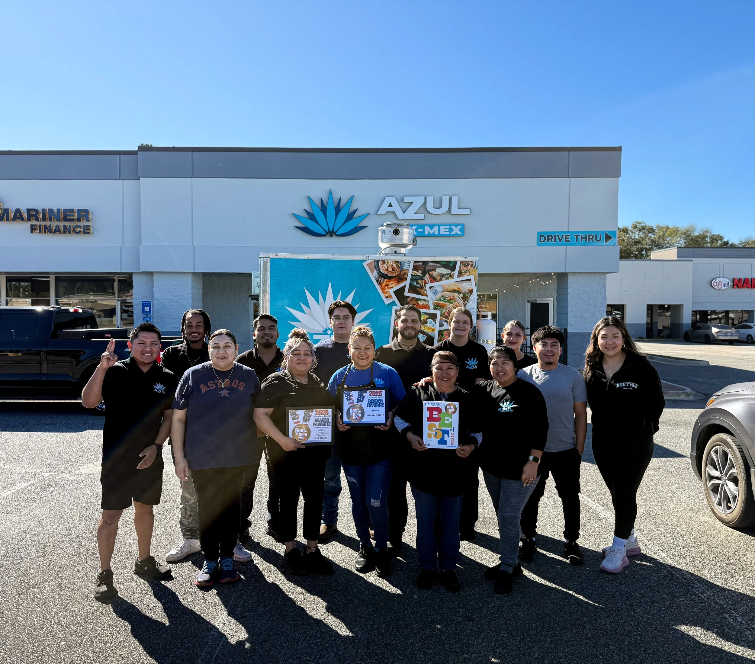 Group of people standing outside AZUL restaurant, some holding awards, with a large blue AZUL sign and food photos in background on sunny day.
