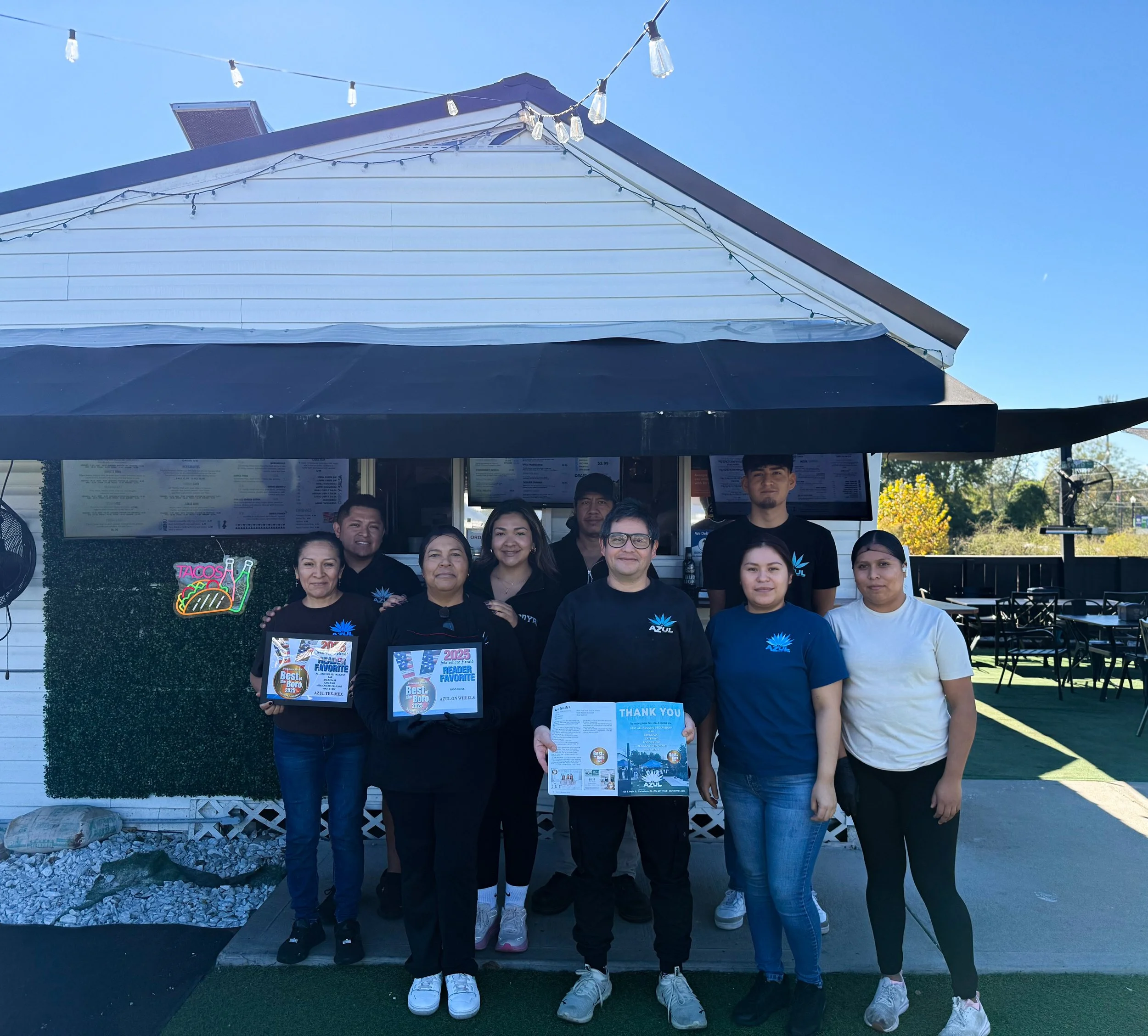 Group of nine people standing outside a restaurant with a sign that says Tacos and three people are holding awards and a flyer, with a clear blue sky in the background.