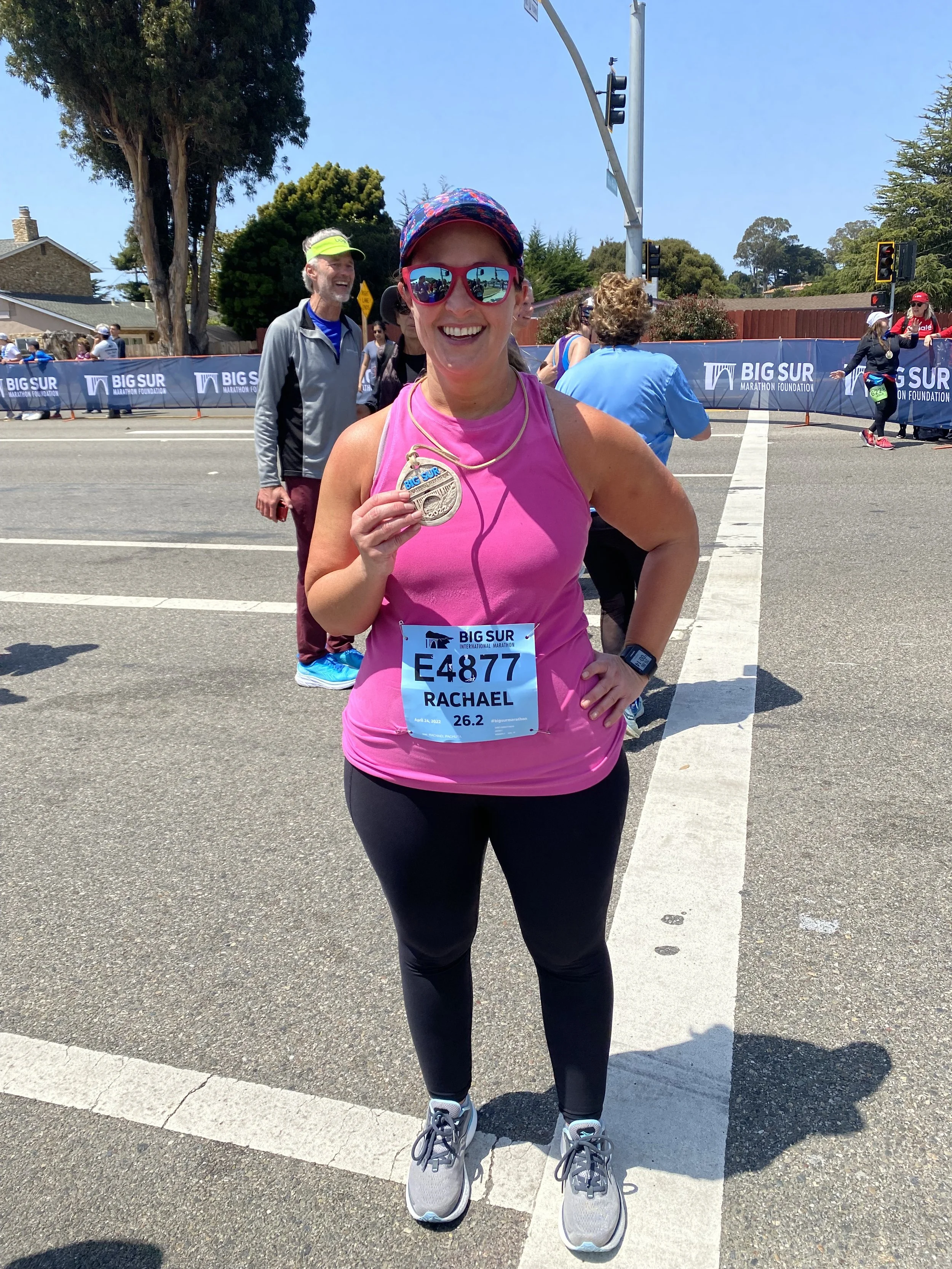 Woman in sunglasses and pink shirt holding a medal with a race bib on