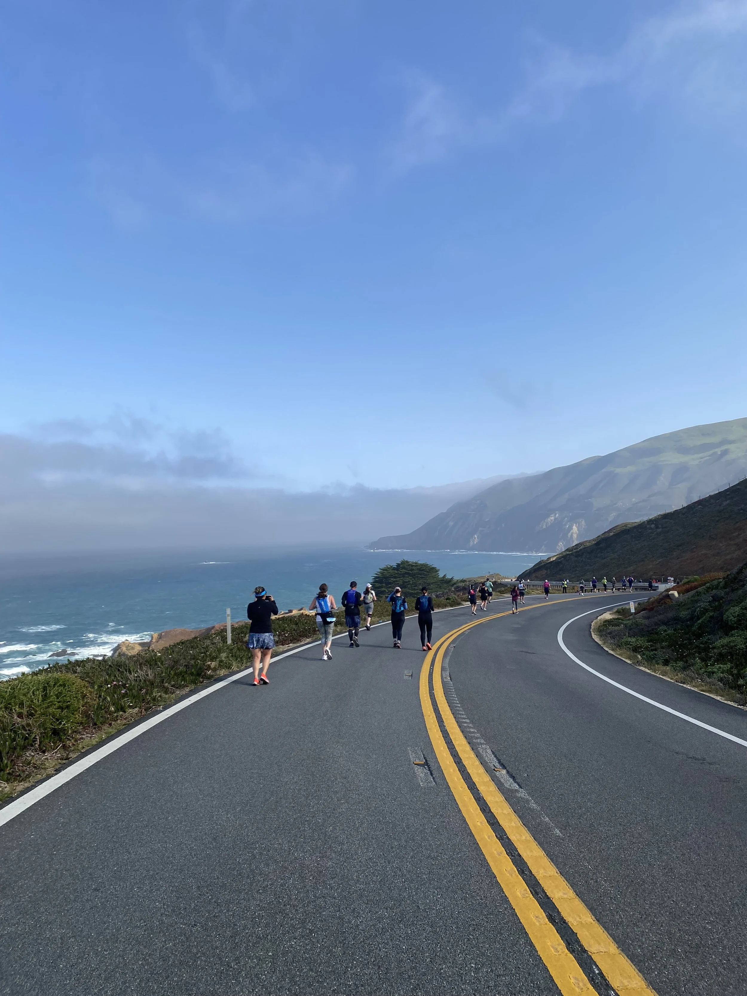 People walking along a winding coastal road with ocean on one side and cliffs on the other, under a partly cloudy sky.