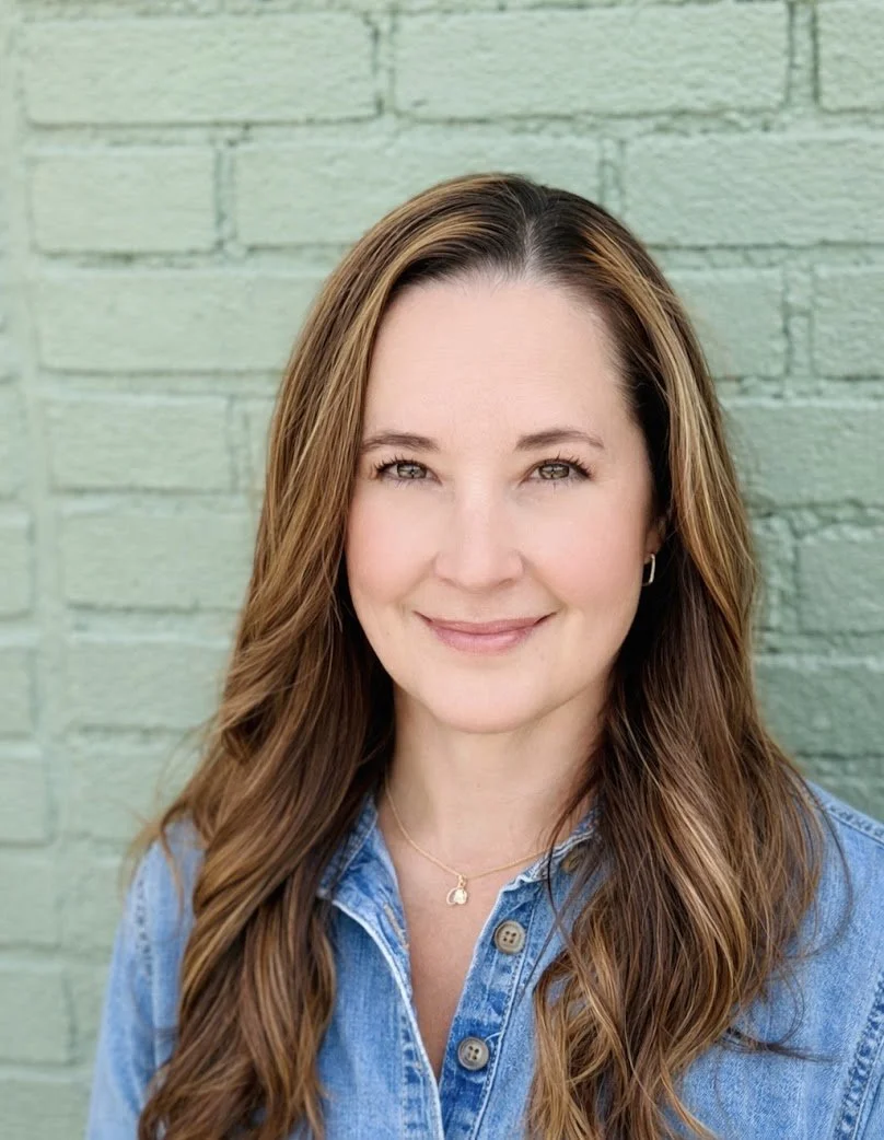 A woman with long wavy brown hair standing in front of a green brick wall, wearing a blue denim shirt and a gold necklace.