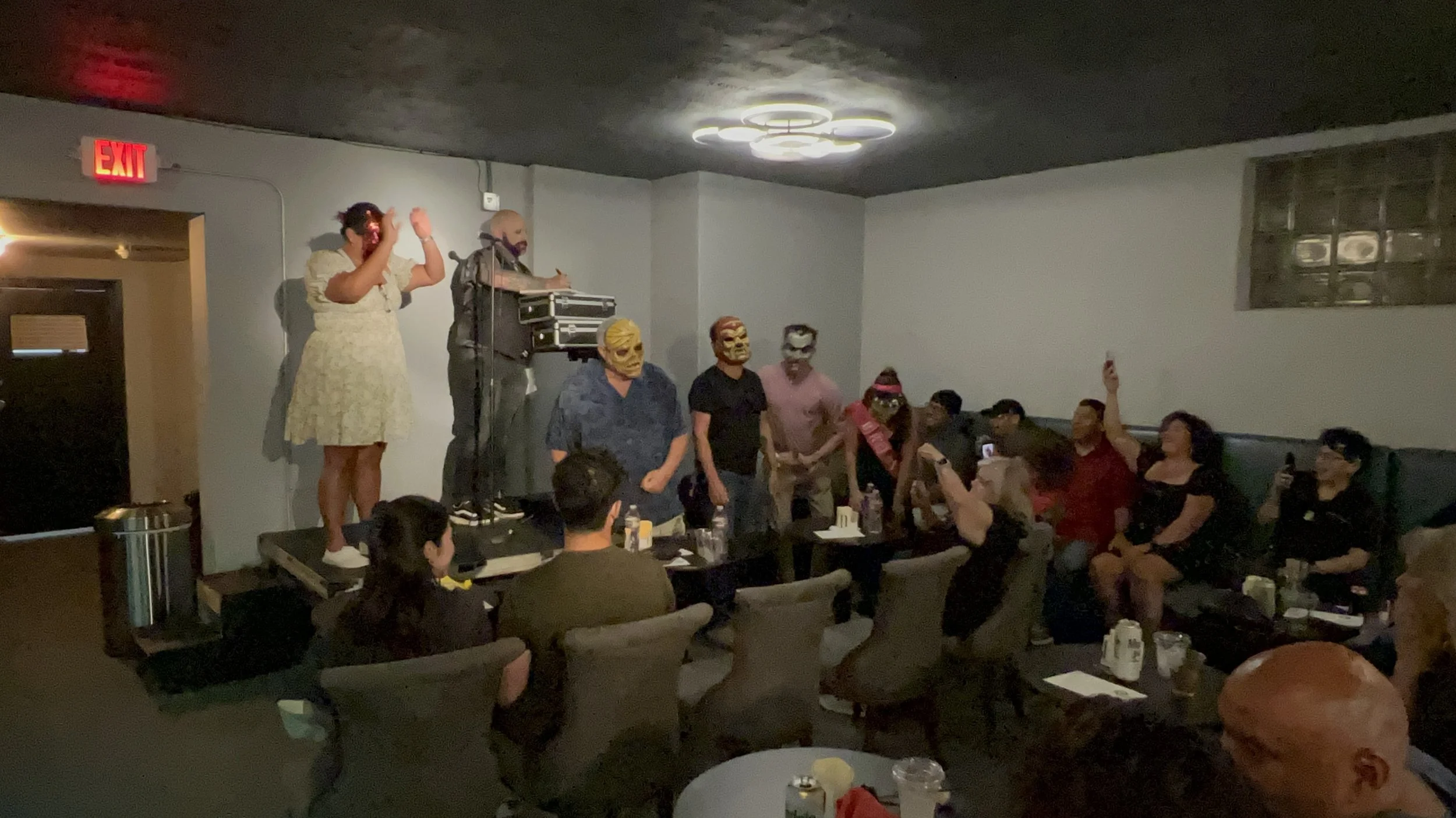 People in masks standing on a small stage in a dimly-lit room, with some taking photos, and audience members seated at tables watching the performance.