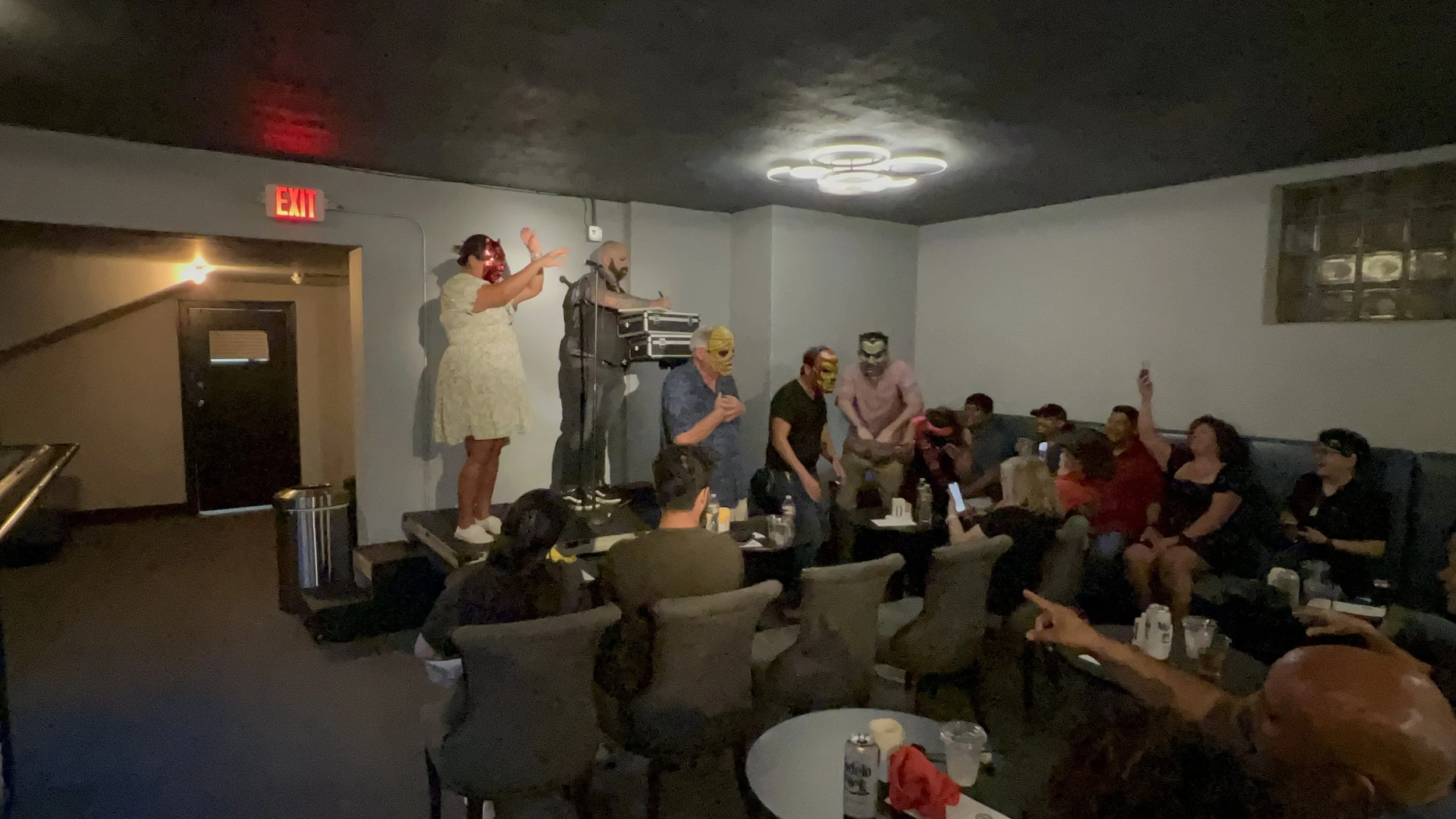 People wearing masks and masks standing on a stage in a dimly lit room, possibly singing or speaking, with a seated audience.