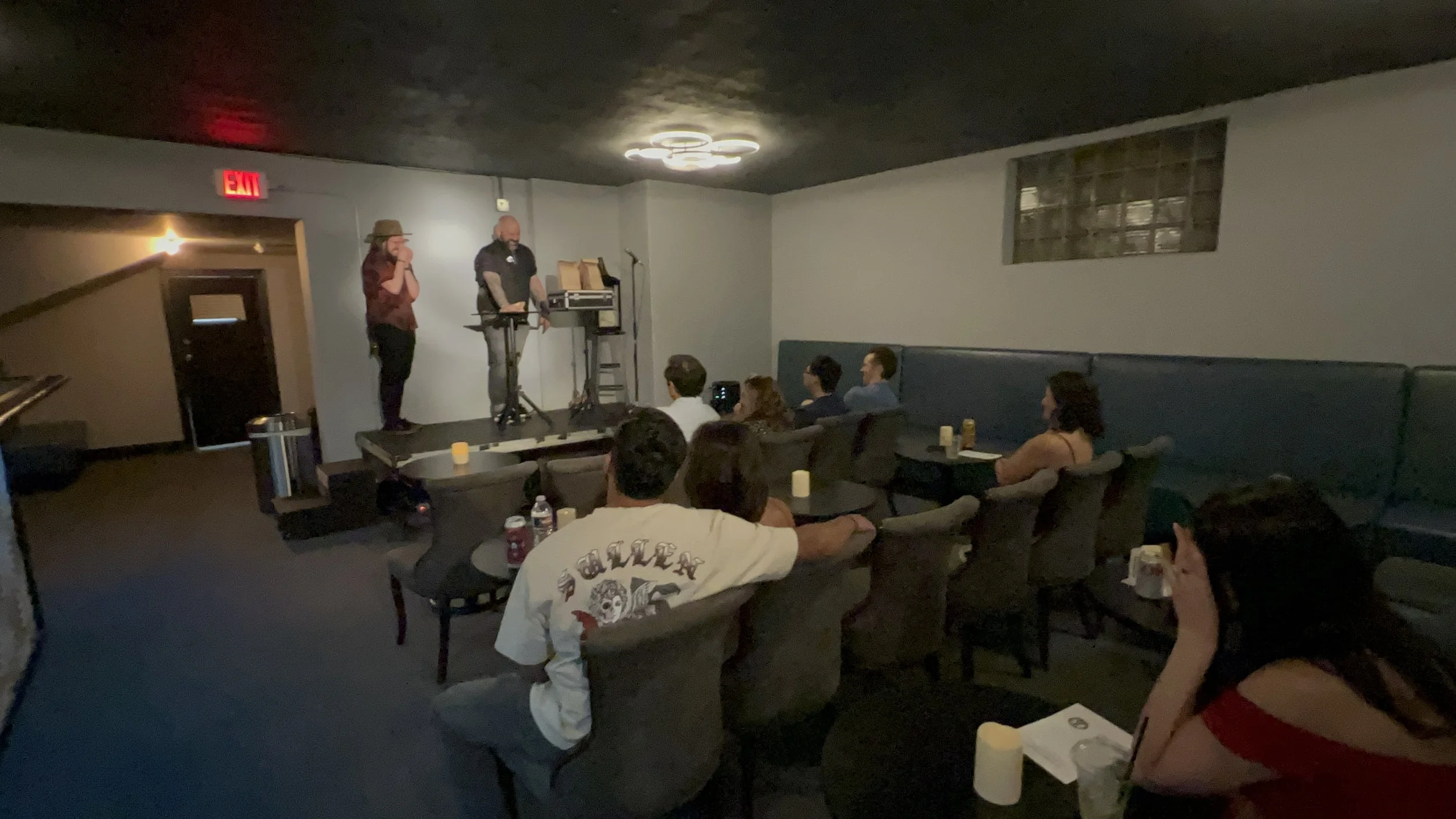 A small comedy show is taking place in a dimly lit room with dark walls. There are two performers on stage: one with long hair and a fedora hat, and the other bald, using a keyboard. An audience of about ten people is seated in chairs and booths, wat