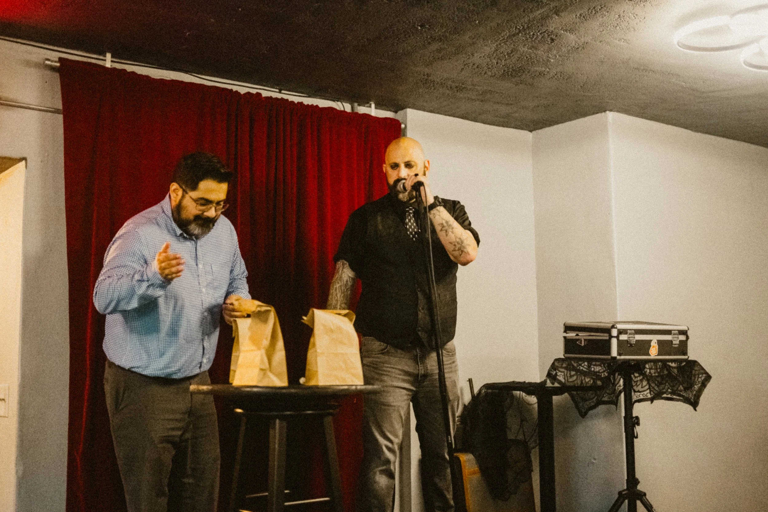 Two men standing on a stage with a red curtain background. One man is holding a microphone and has tattoos on his arms, while the other man is looking down at paper bags on a small table.
