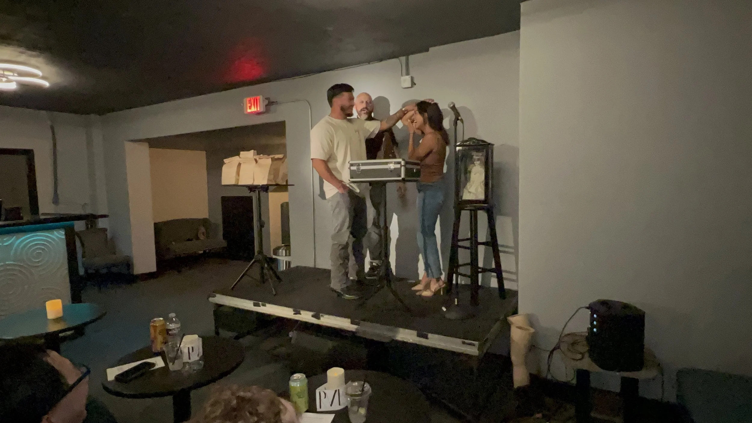 A group of people on a small stage in a dimly lit venue. One man appears to be performing as a magician or illusionist with a woman as his assistant, while another man watches. There are tables with drinks and items around the room, and a microphone 