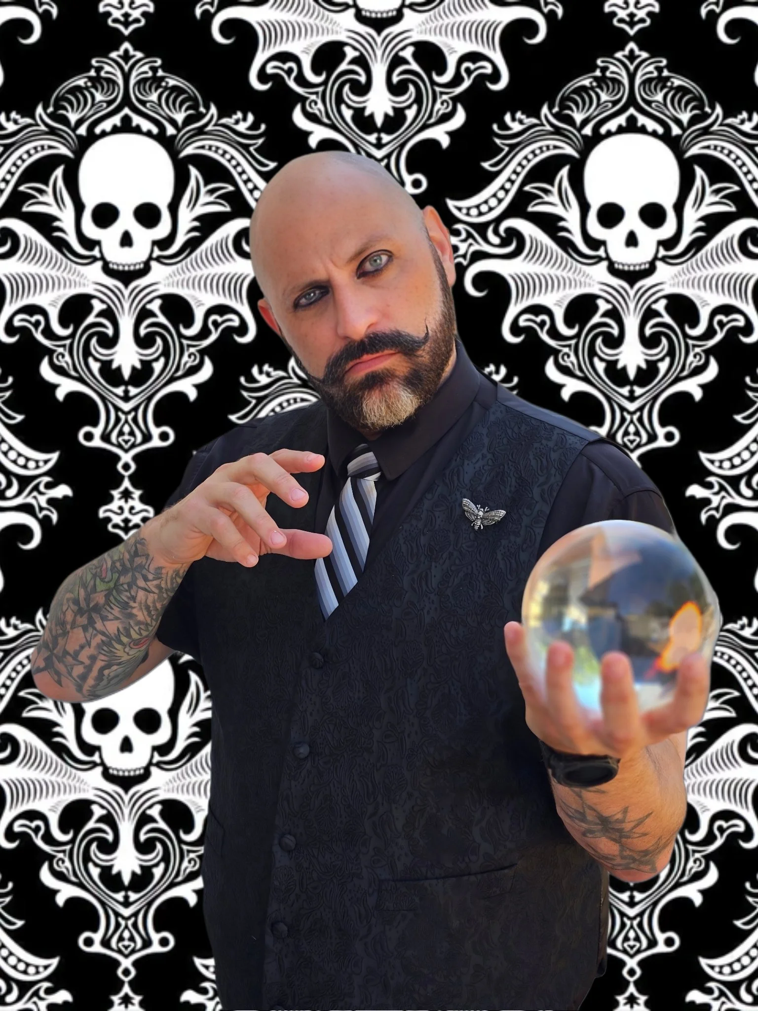 A magician with a beard and tattoos holding a crystal ball, dressed in a dark vest and tie, with a black and white skull-patterned background.