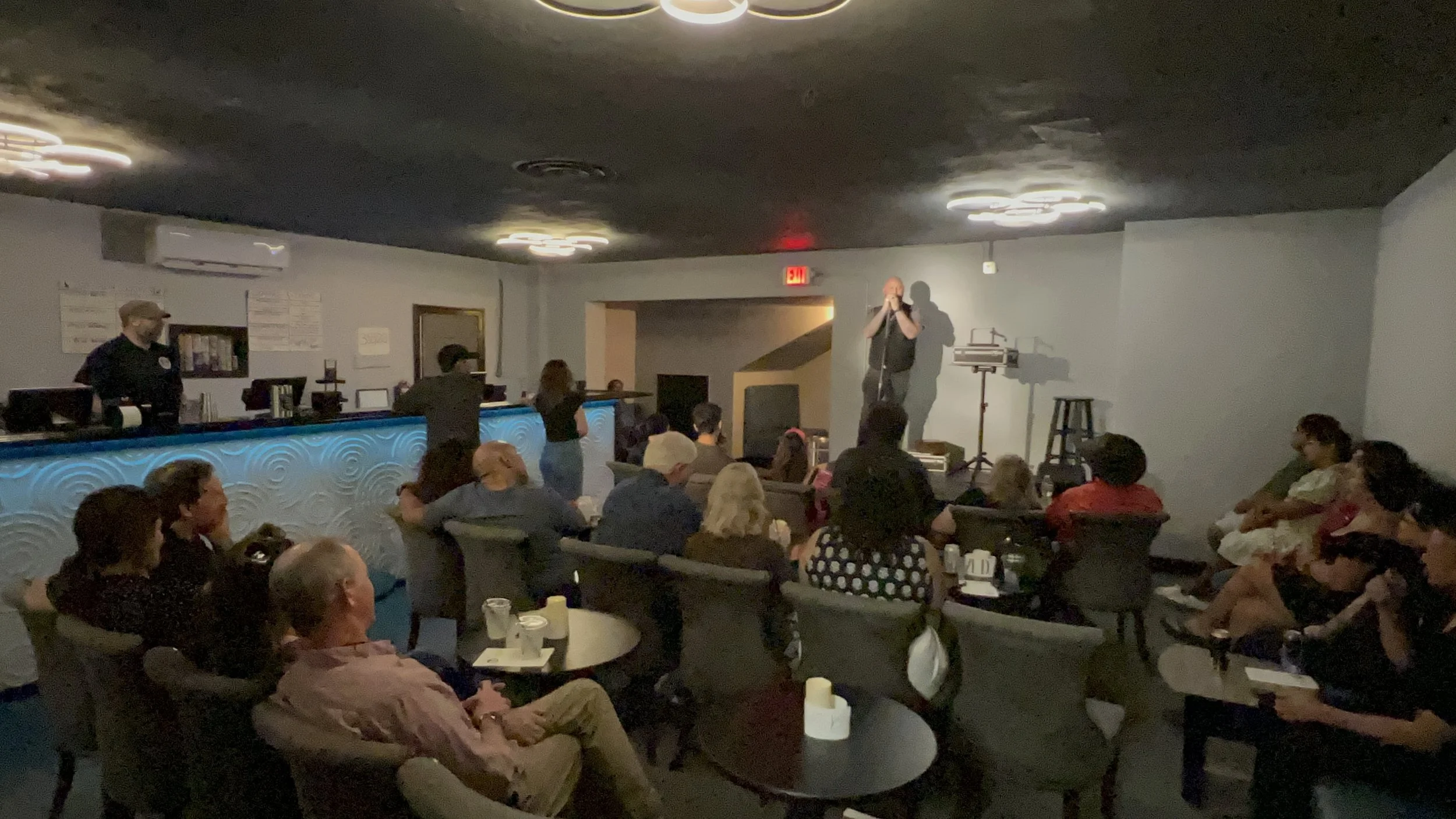 A comedy club or lounge with a group of people seated and listening to a performer on stage. The performer is a bald man with a black t-shirt, standing and speaking into a microphone. The audience is seated in chairs around small tables, some with dr