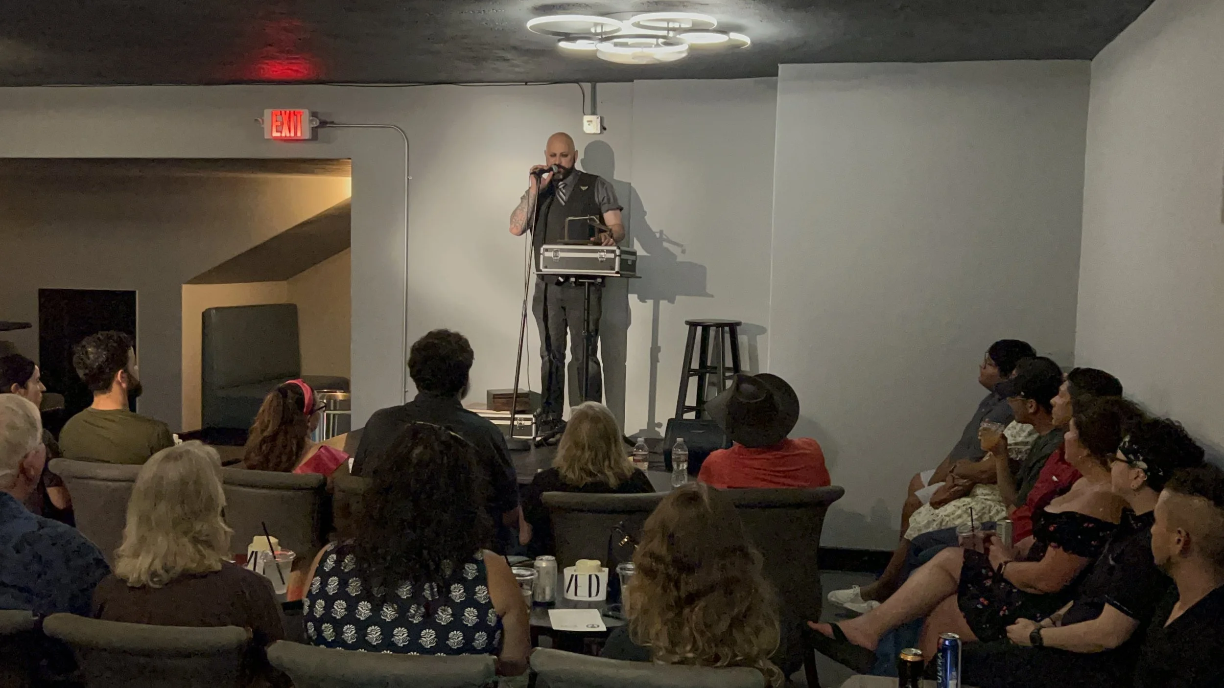 A man on stage performing with a microphone and electronic equipment, while an audience watches attentively in a dimly lit room.
