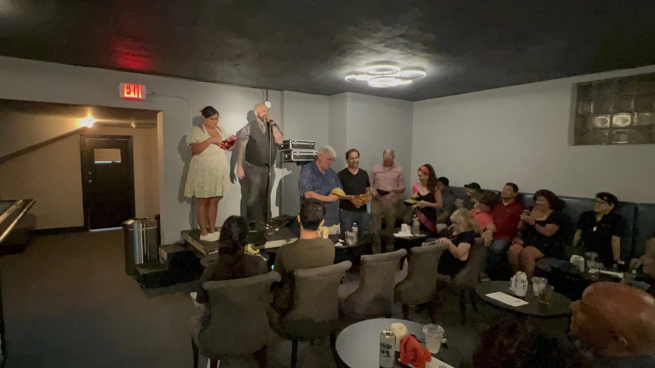 People gathered in a dimly lit room, some sitting around tables and others standing on a small stage. There are two individuals on stage, one with a microphone, and the audience appears to be listening to a speech or presentation.