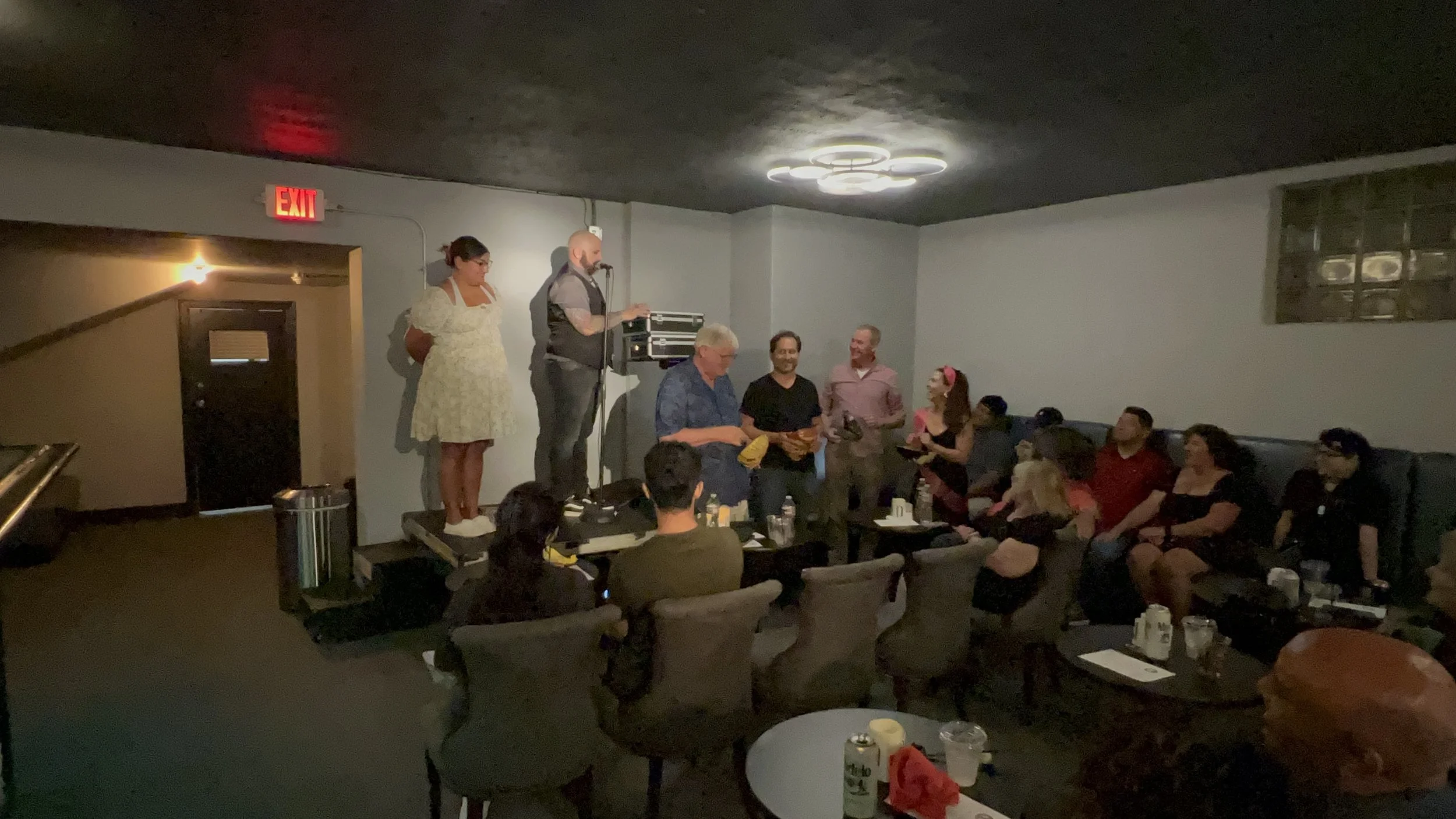 Group of people gathered at a karaoke night with some on stage singing and others sitting on couches watching and smiling in a dimly lit room.