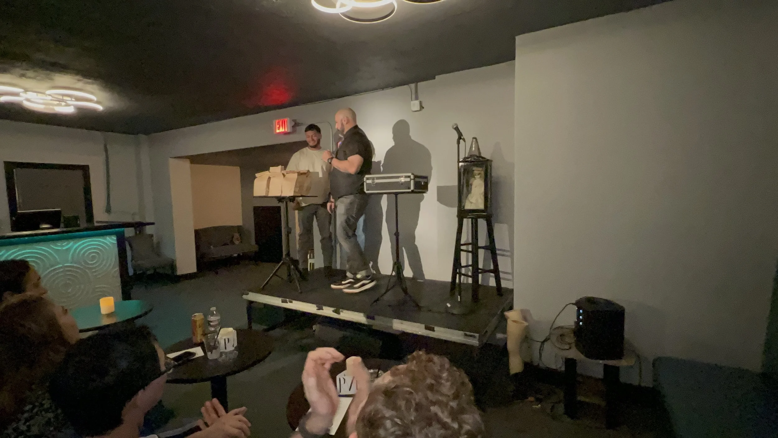 Two men stand on a small stage, one speaking into a microphone while the other looks at him. There is audio equipment on the stage and an audience seated in front, watching the performance. The setting appears to be a comedy or open mic night in a di