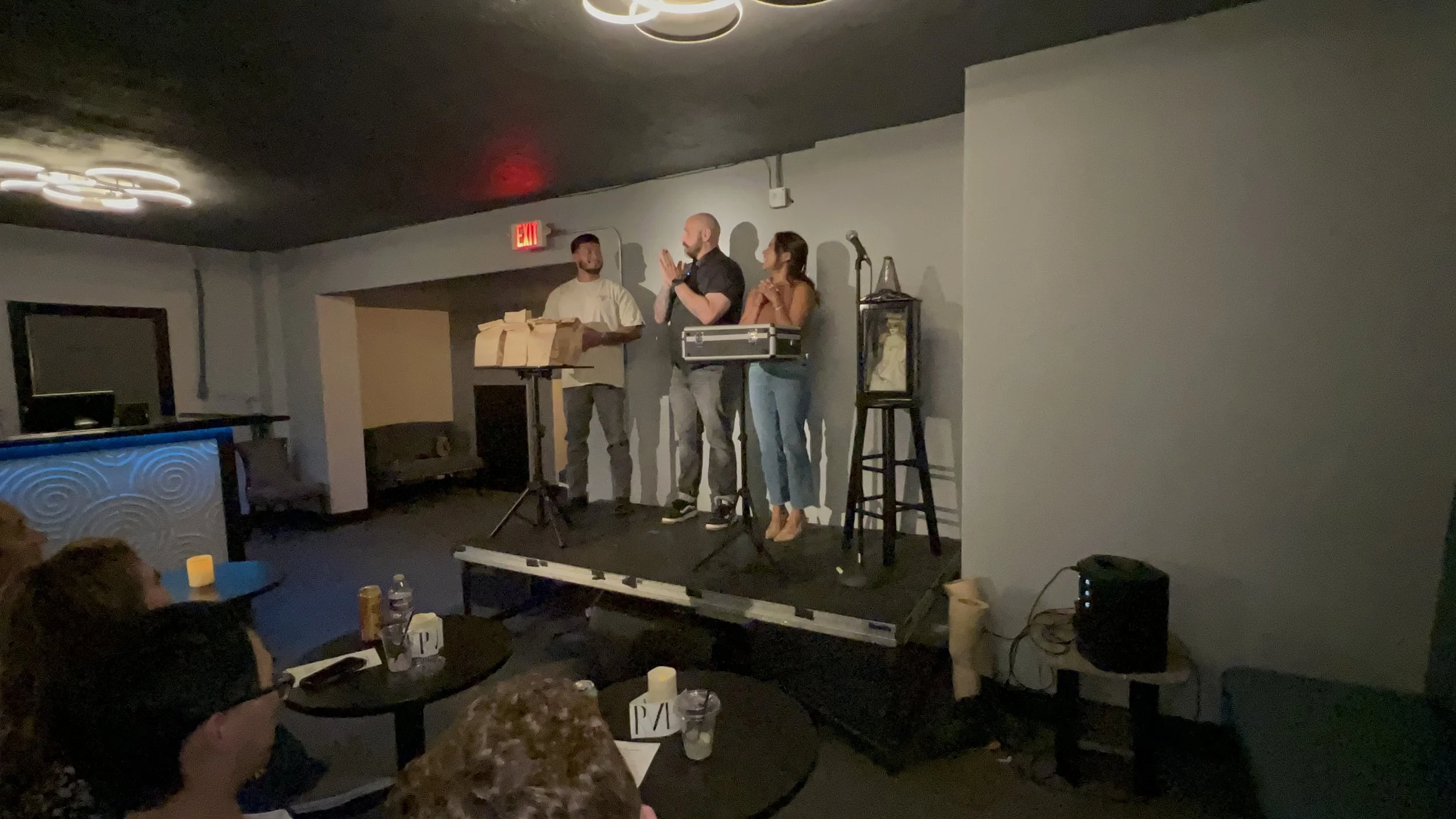 People on stage performing in a dimly lit venue, with audience seated at tables watching.