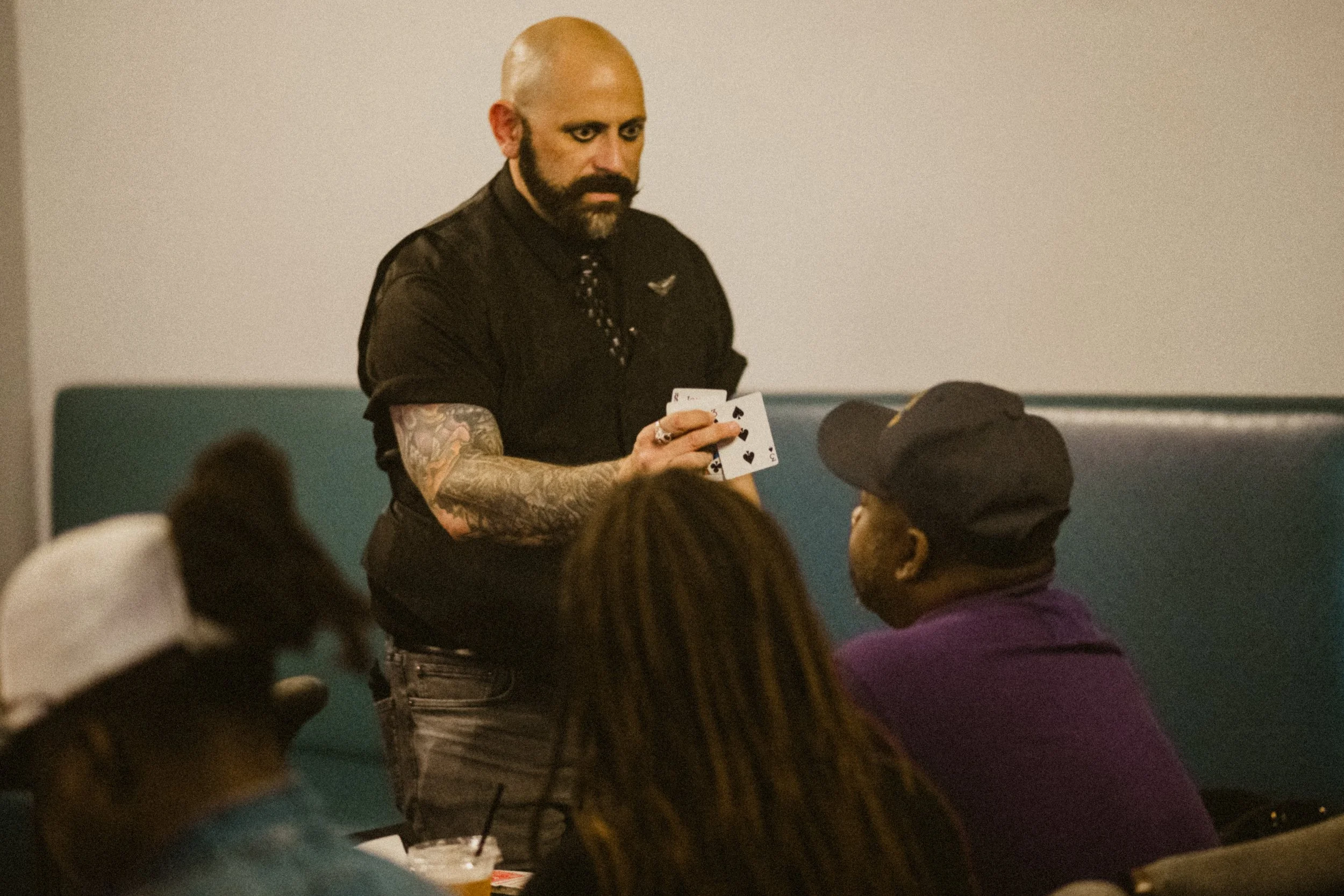 A magician with tattoos, a beard, and makeup holding playing cards and performing a magic trick in front of seated audience members in a dimly lit room.