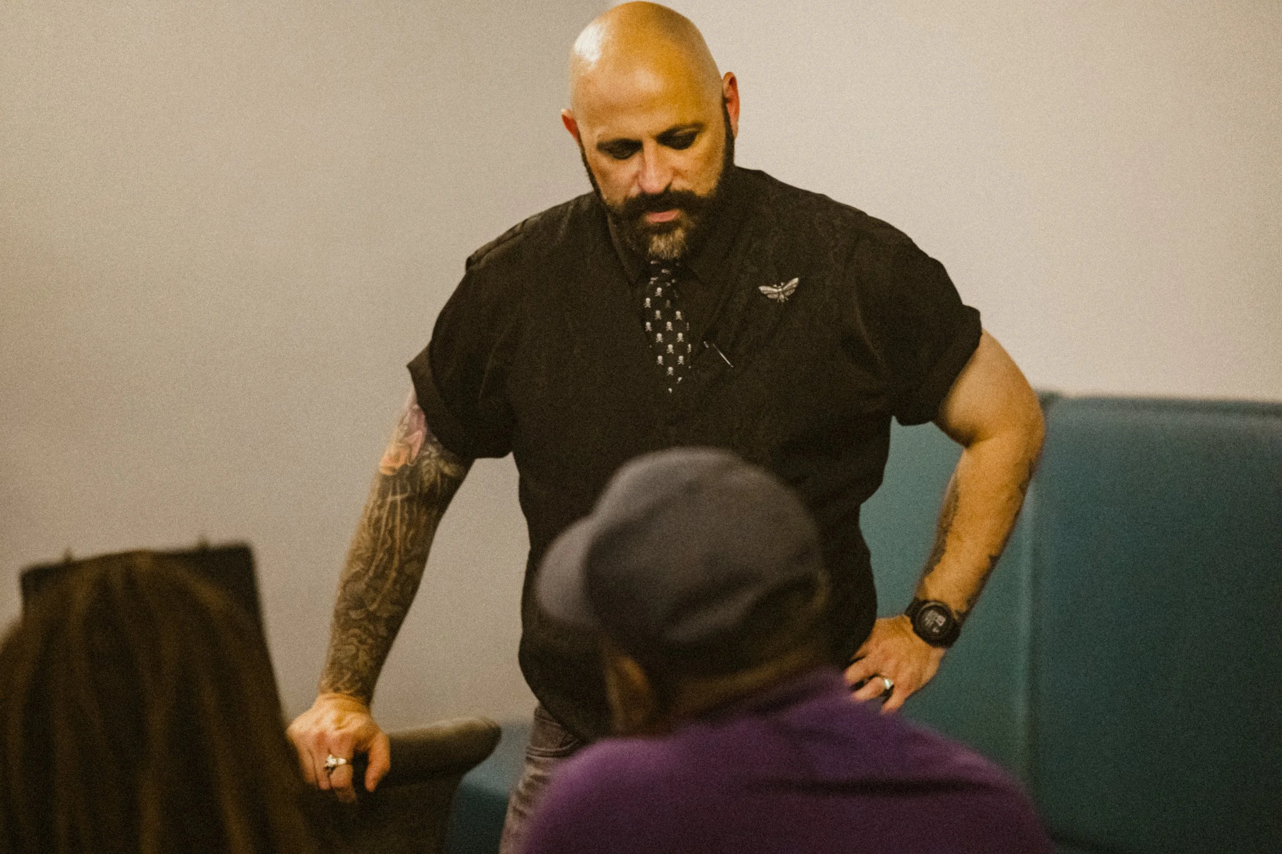 A man with a shaved head and beard, dressed in black with tattoos on his arms, is leaning on a table and speaking to seated individuals in a room with plain walls.