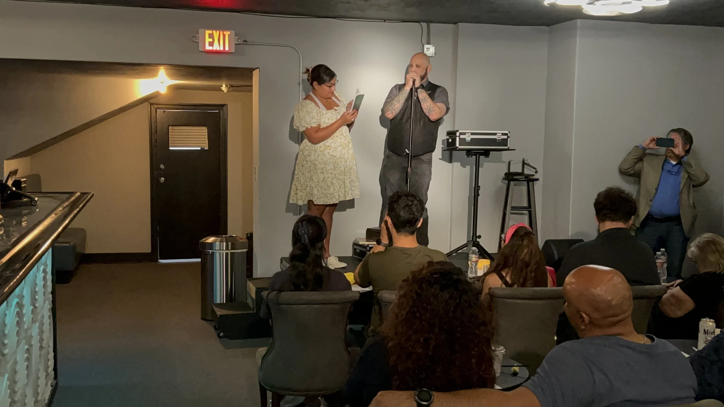 A woman and a man on a stage, the woman is reading from a paper while the man speaks into a microphone, with an audience seated in front. An observer is taking a photo or video, standing against the wall.