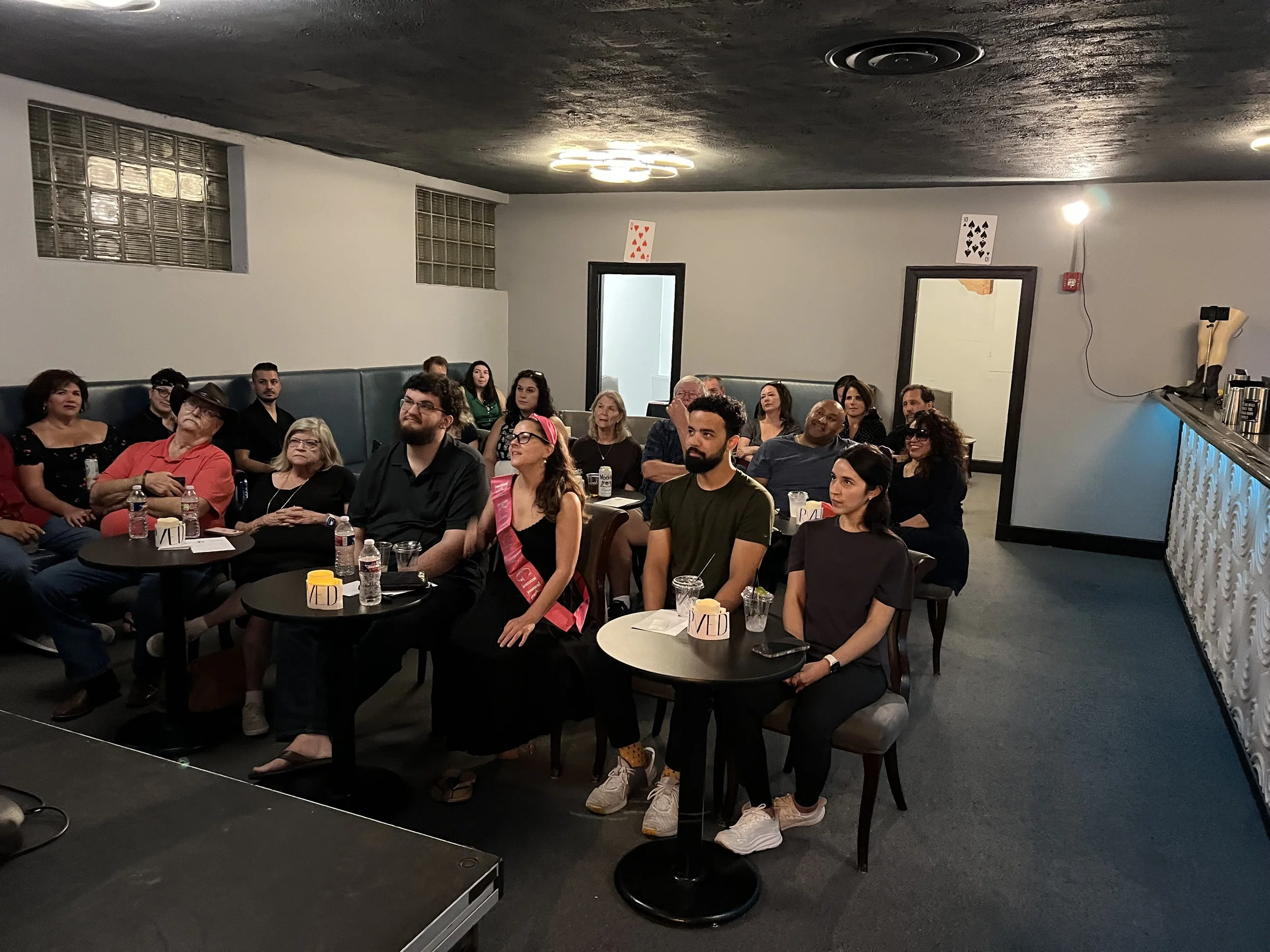 Group of people attending a presentation or performance in a dimly lit room, sitting at tables with drinks, some watching attentively.
