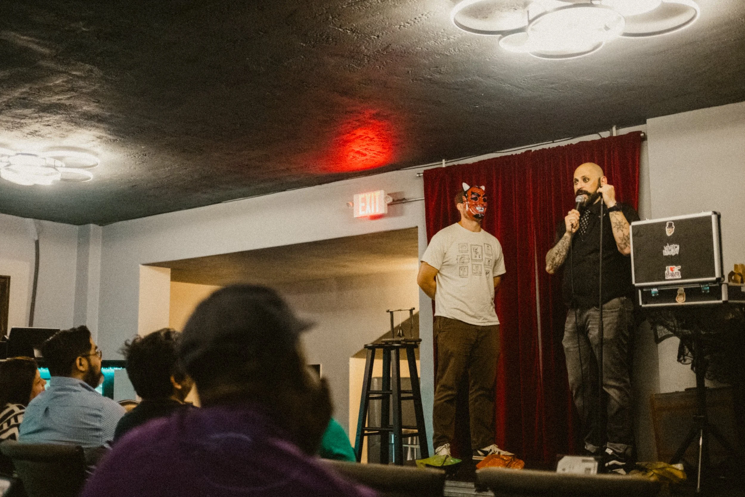 A magician with a tattooed arm and wearing a black shirt is speaking into a microphone on stage. Next to him stands a man wearing a devil mask.  The audience is seated and watching the performance.