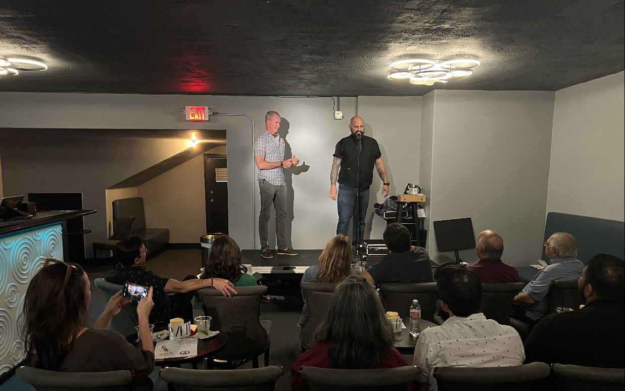 A comedy performance or speech on a small stage with two men, one with a microphone, addressing an audience in a dimly lit room with modern ceiling lights.